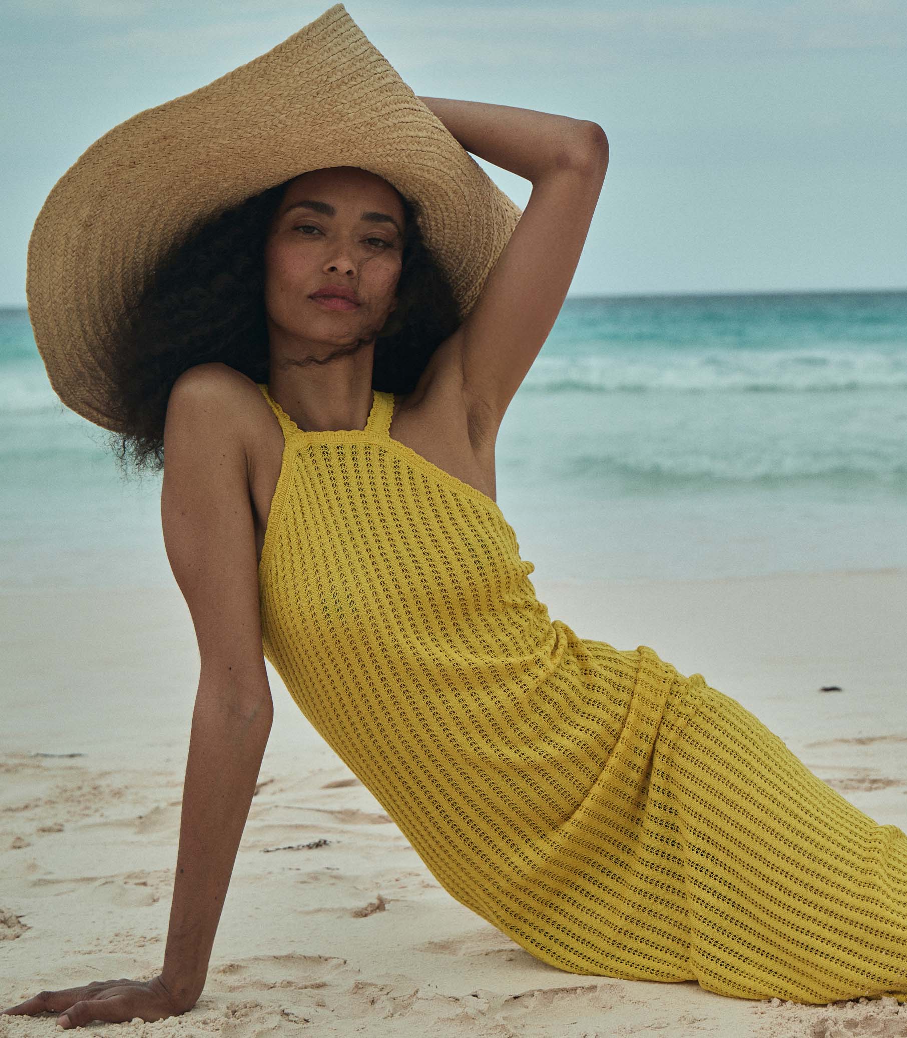 Anais is wearing a sleeveless yellow knit maxi dress with a sun hat on the beach. 1
