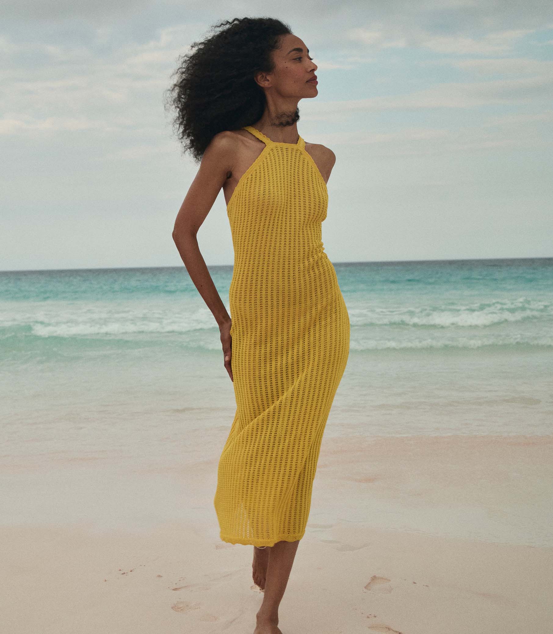 Anais is wearing a sleeveless yellow knit maxi dress with a sun hat on the beach. 5
