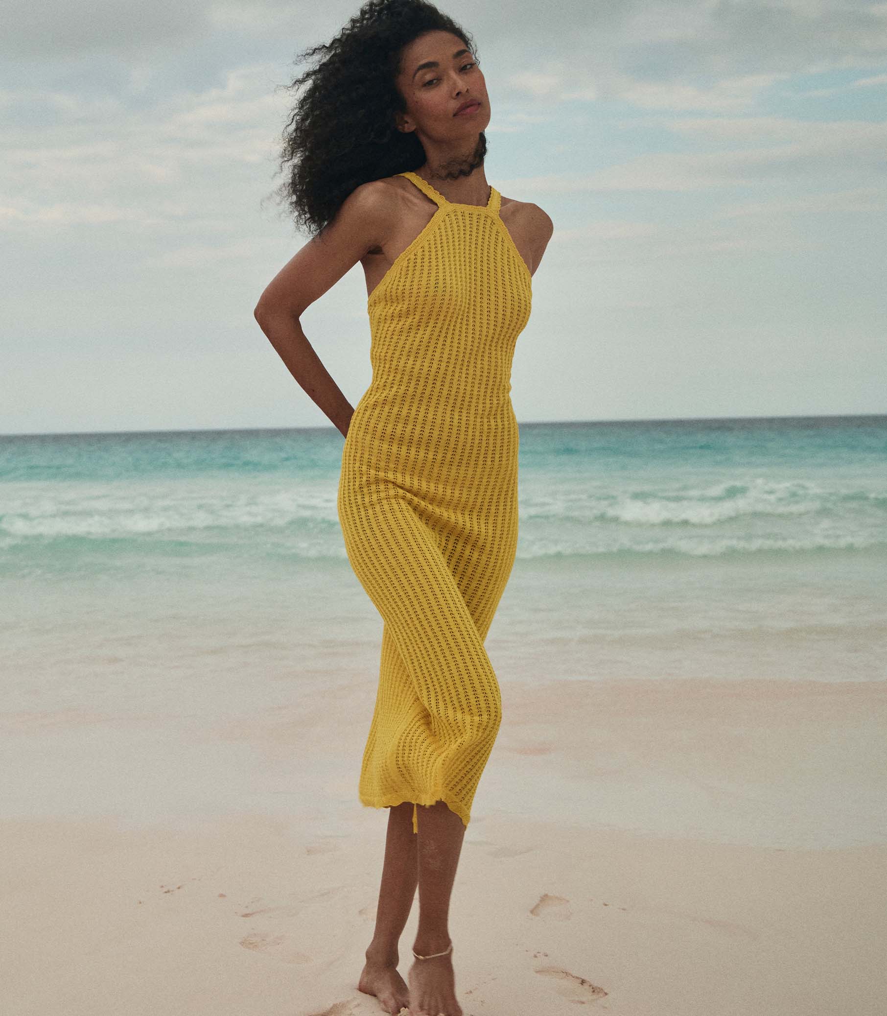 Anais is wearing a sleeveless yellow knit maxi dress with a sun hat on the beach. 4
