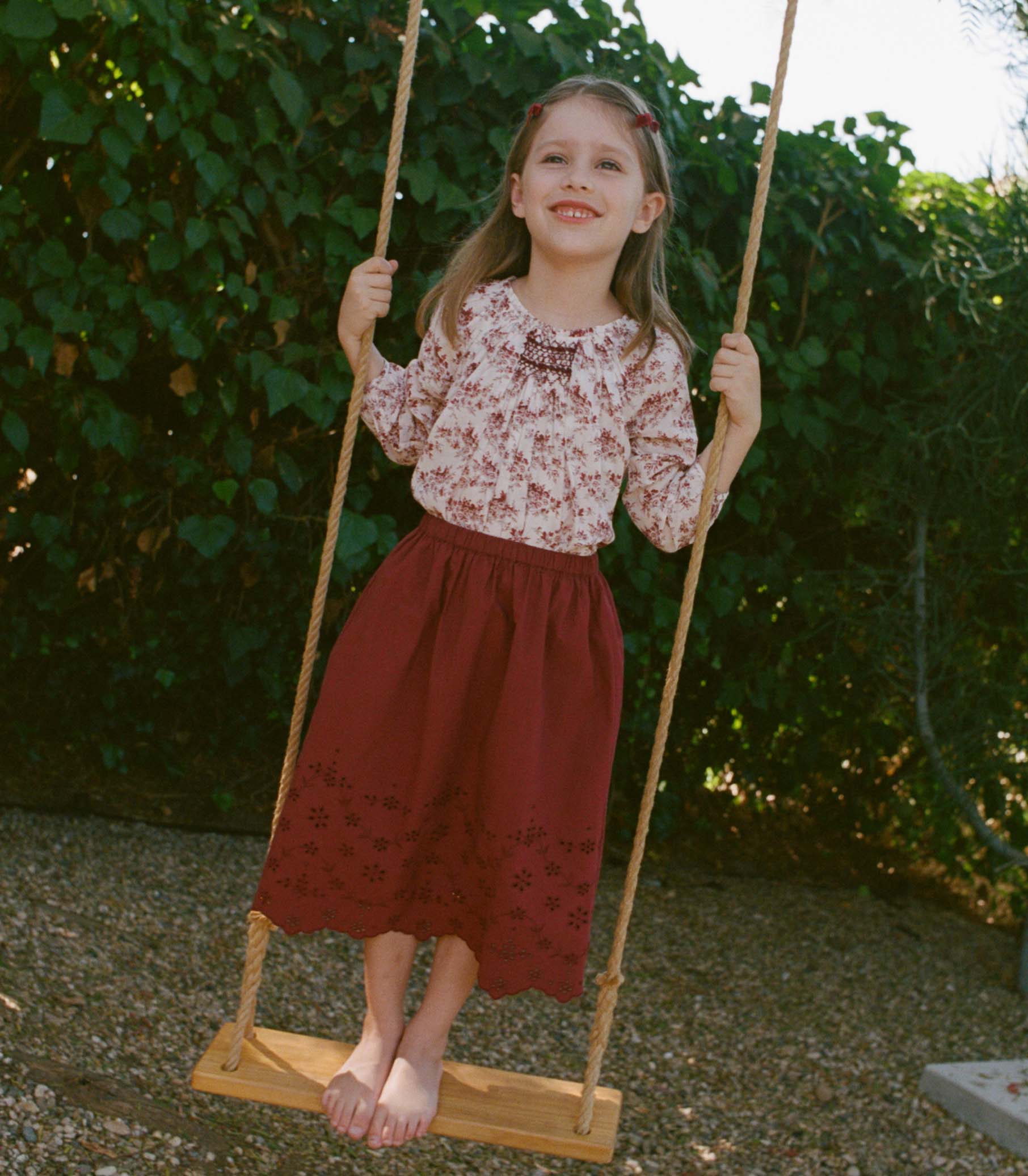 Young girl in a floral top and red skirt sitting on a wooden swing with greenery in the background. 1
