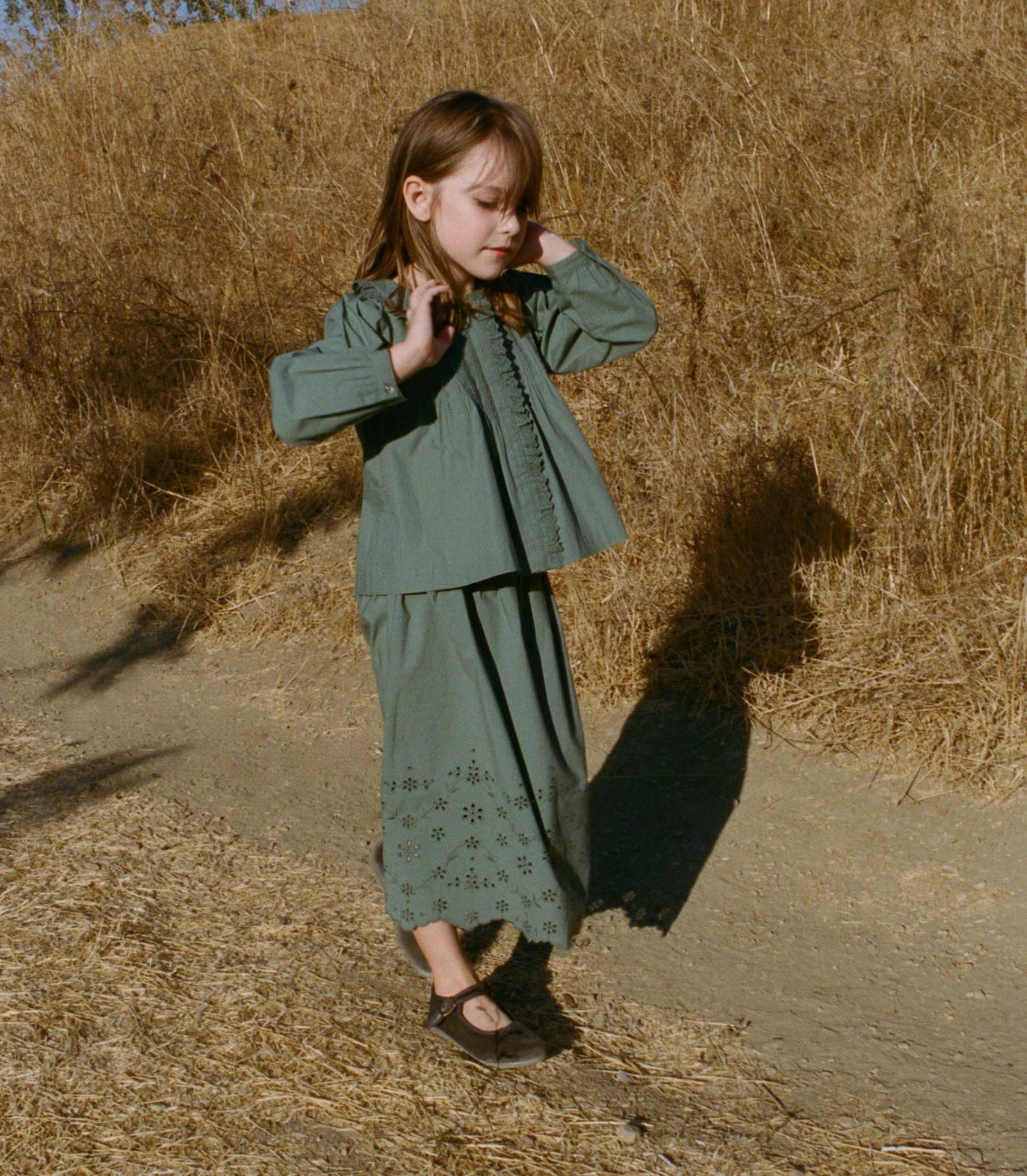 Young girl in a teal top and matching skirt, standing in a field of dry grass. 1
