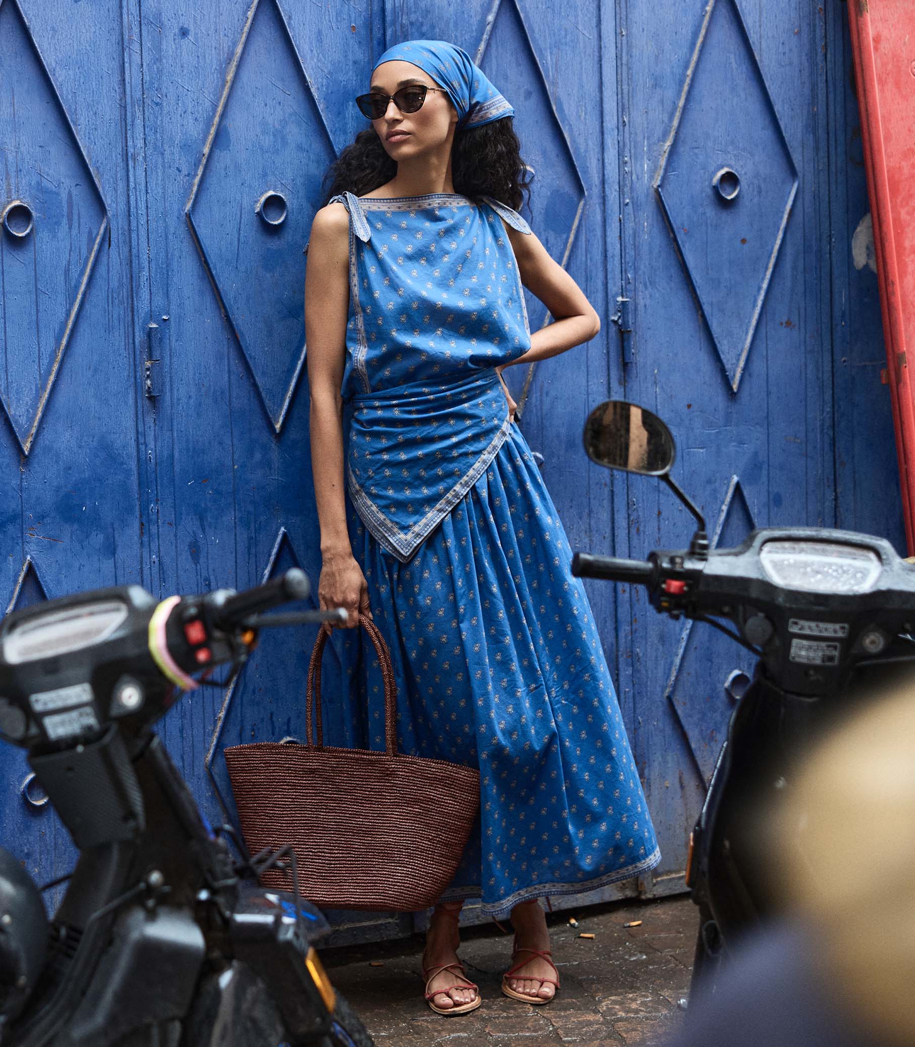 Woman in a blue paisley skirt and matching top standing next to a motorcycle with a blue wall in the background 1
