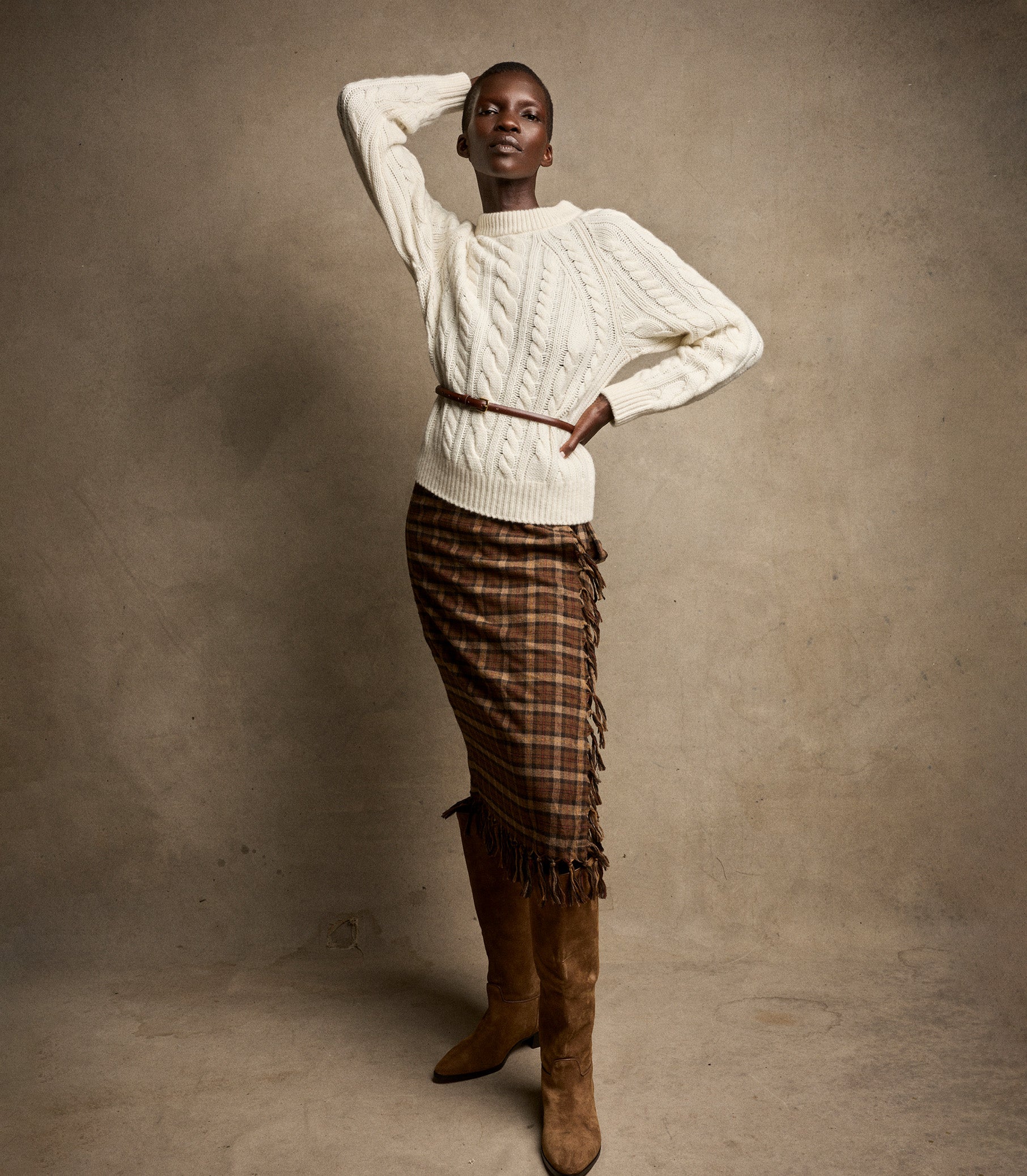 Woman wearing a white cable knit sweater and plaid skirt with brown knee-high boots against a beige background 2
