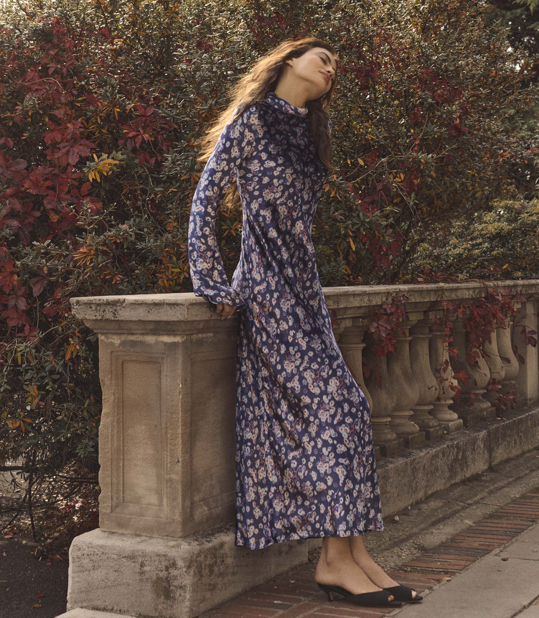 Woman in a purple floral dress standing against a stone wall with foliage in the background