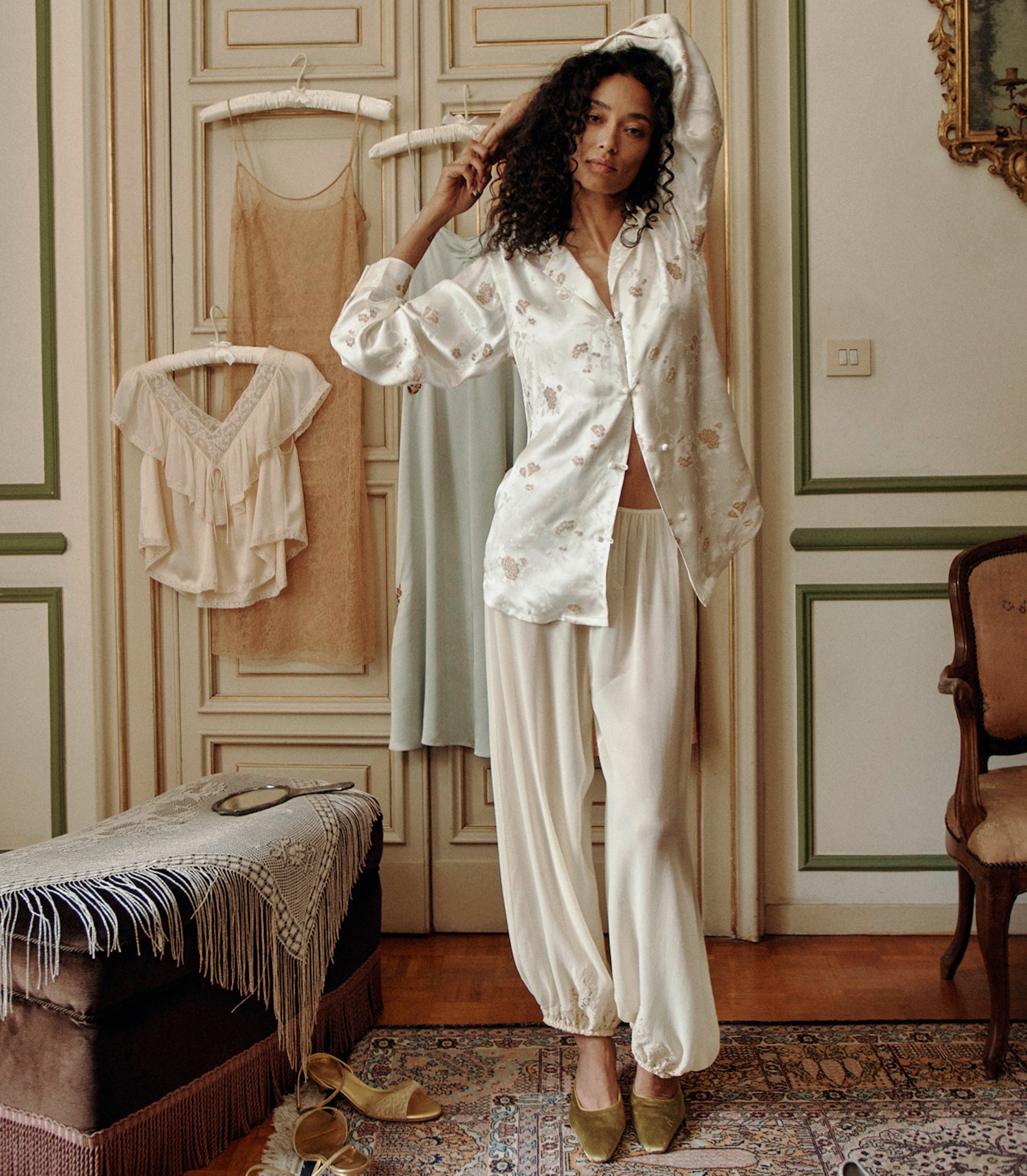 A woman stands in a decorated room wearing a white satin blouse with floral embroidery, consisting of a long-sleeve button-up shirt and loose pants. Other garments hang on hangers on a door behind her.