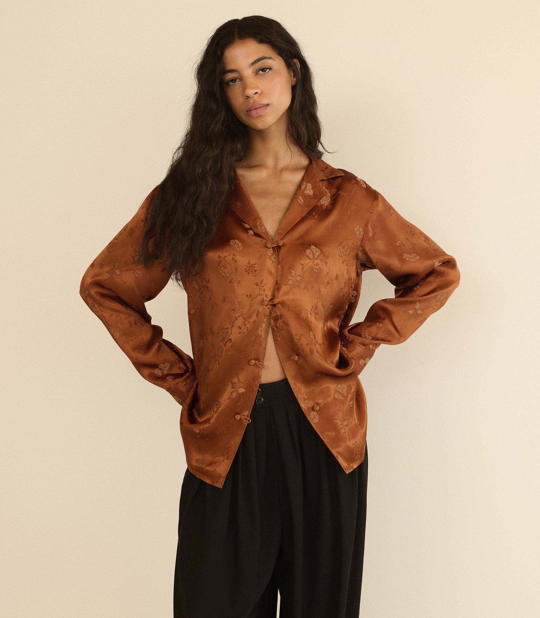 A woman modeling a long-sleeve satin-like button-front blouse in a warm rust brown with a subtle tonal floral jacquard pattern, paired with black pants, against a plain beige studio background.