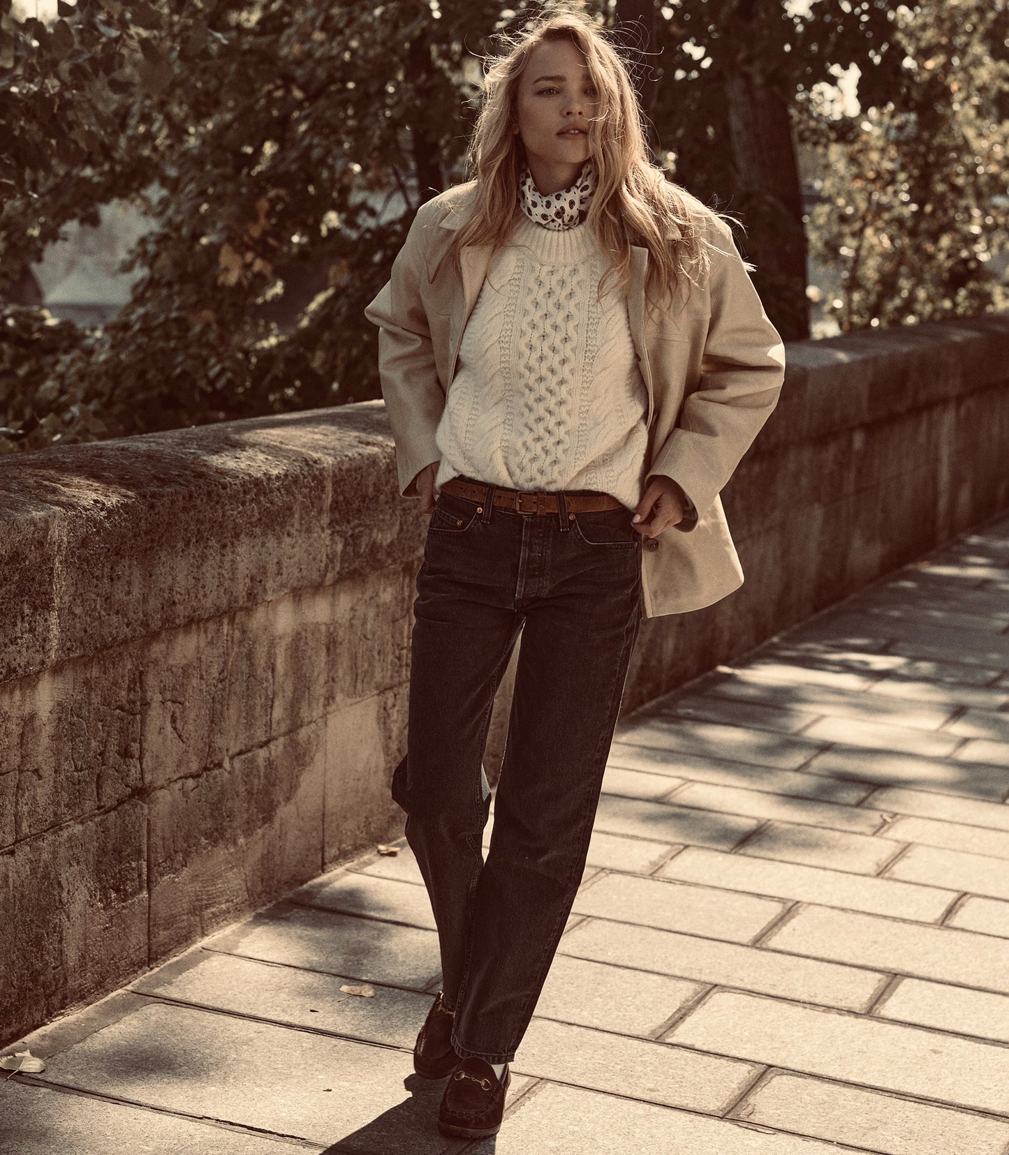 A woman stands on a stone walkway outdoors wearing a beige trench coat over a cream cable-knit sweater, dark jeans, and brown shoes.