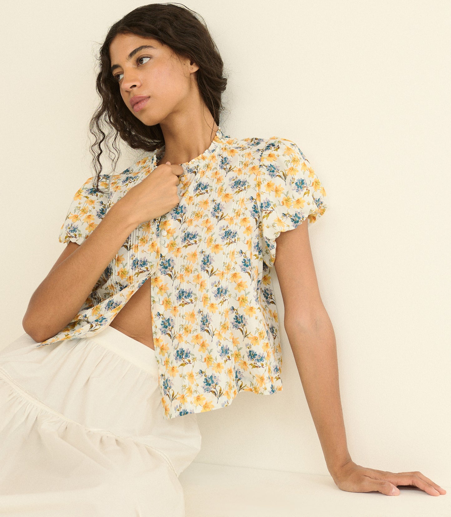 A woman wearing a short-sleeve yellow and blue floral print blouse with puff sleeves and a high neck, paired with a white ruffled skirt, posed against a plain cream background.