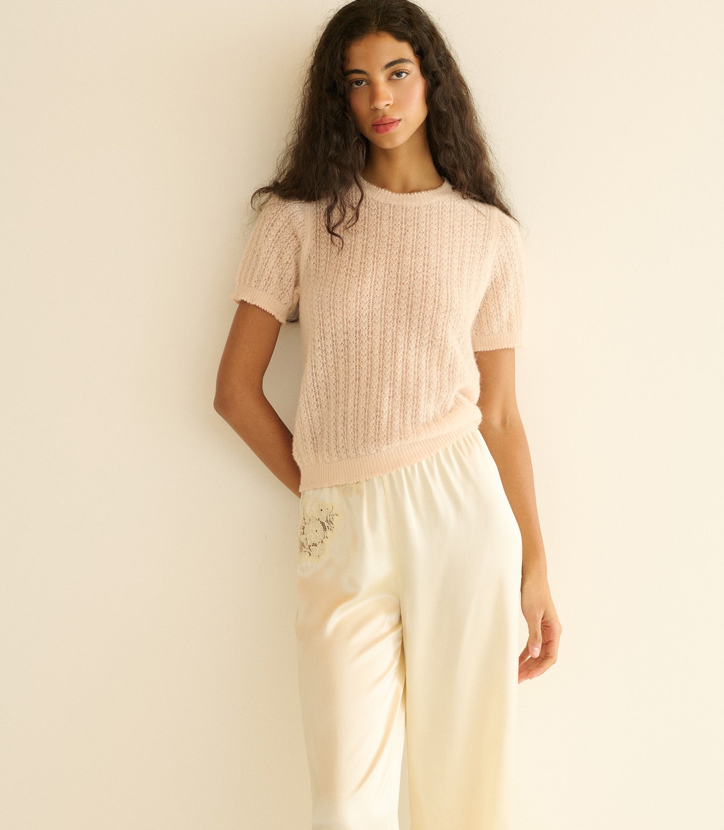 Woman wearing a light pink short sleeve sweater and white pants against a plain background