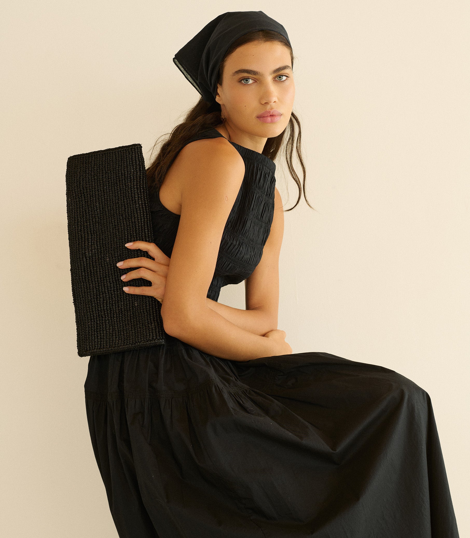 Woman in a black dress holding a black clutch against a beige background 1
