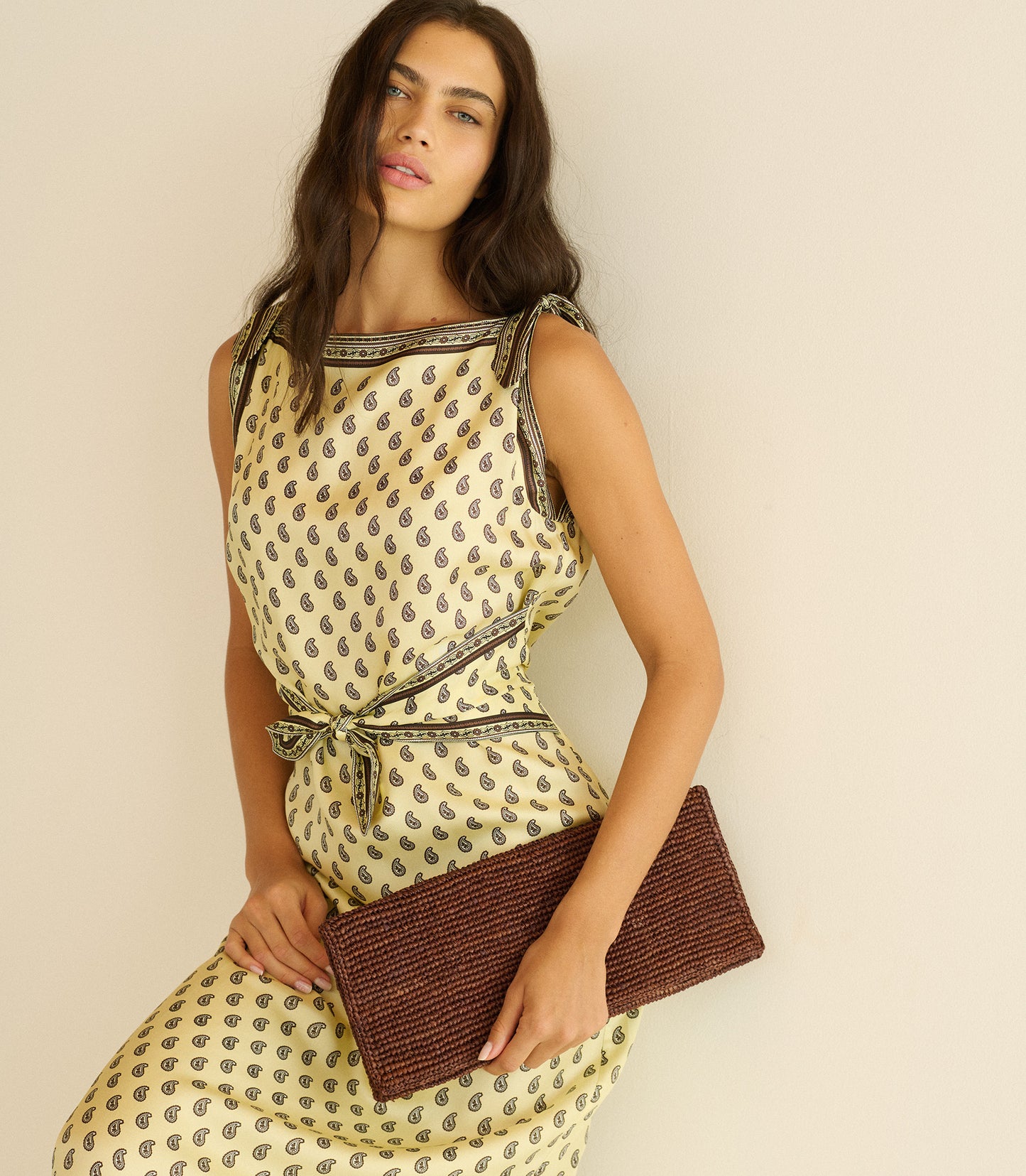 Woman wearing a patterned dress holding a reddish brown clutch against a beige background