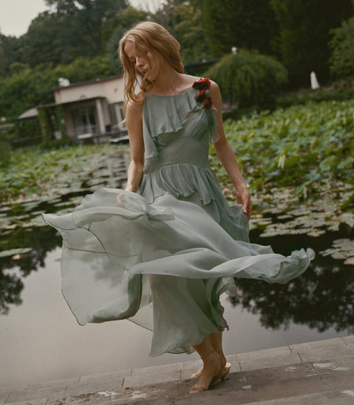 A woman in a flowing pale sage green midi dress walks barefoot beside a pond with lily pads, holding a small red rose.