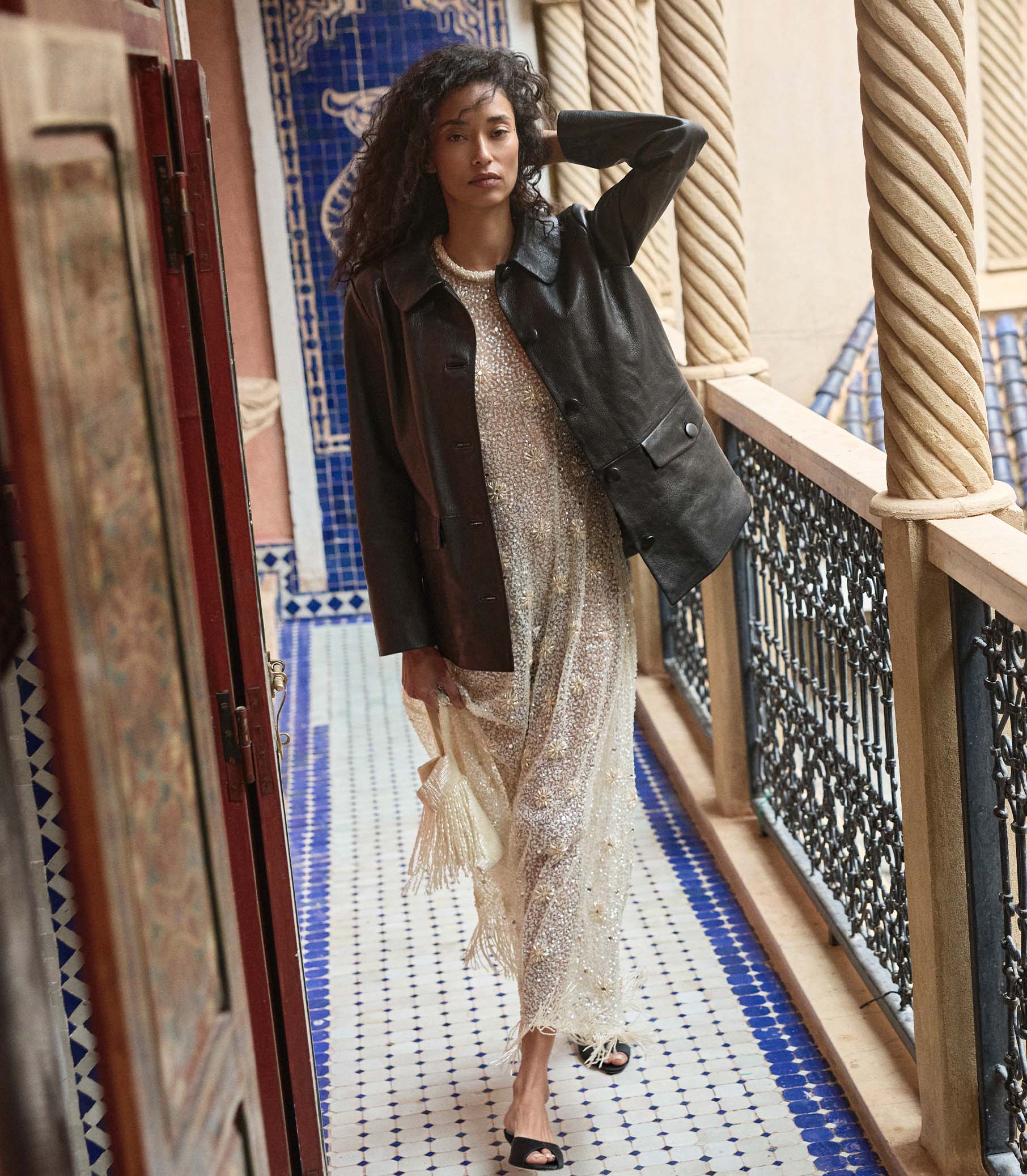 Woman in a stylish dress and black leather jacket standing on a balcony with decorative tiles.