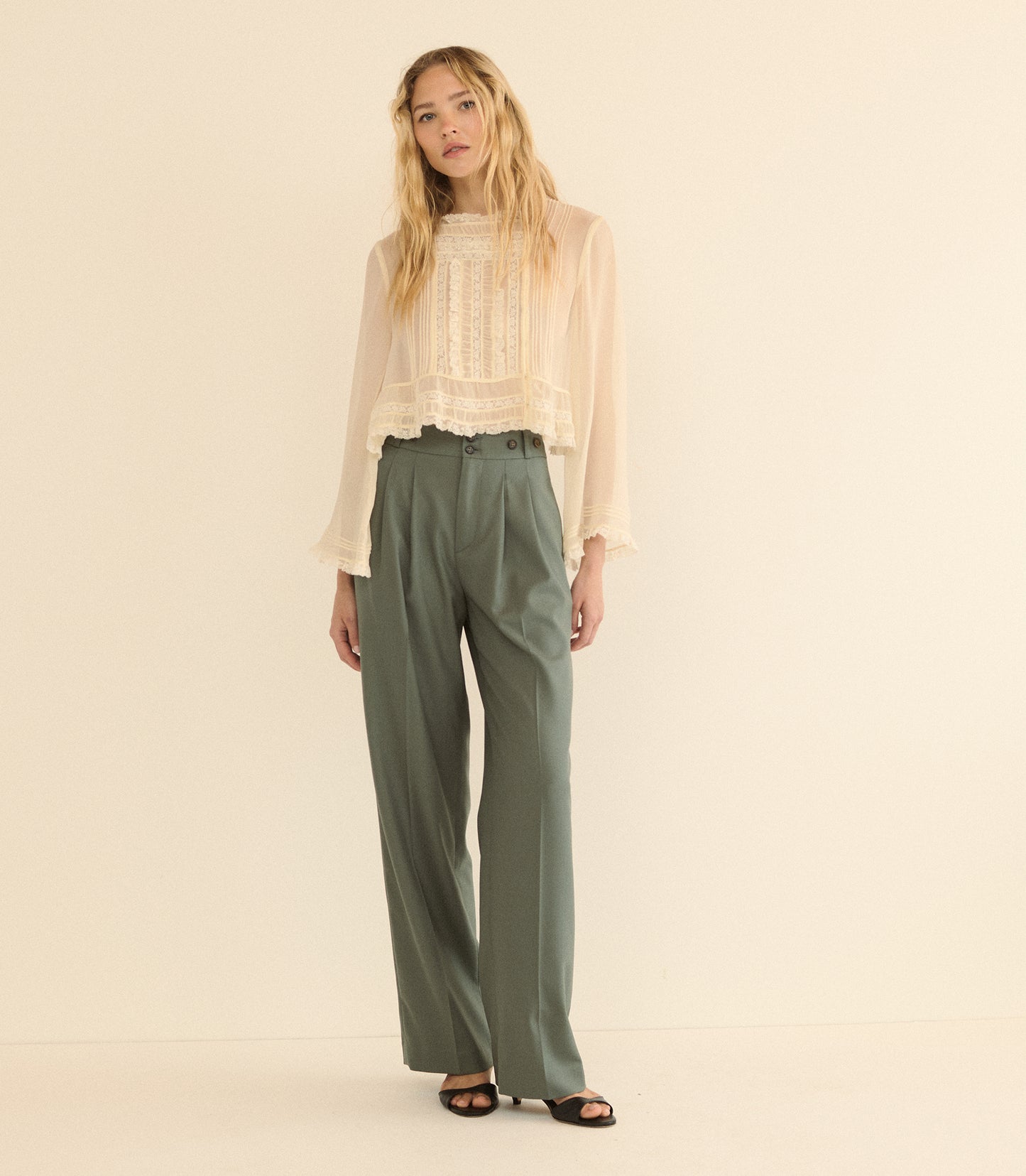 A woman stands against a plain warm cream studio background wearing a sheer long-sleeve cream blouse with a lace front and high-waisted olive green wide-leg trousers.