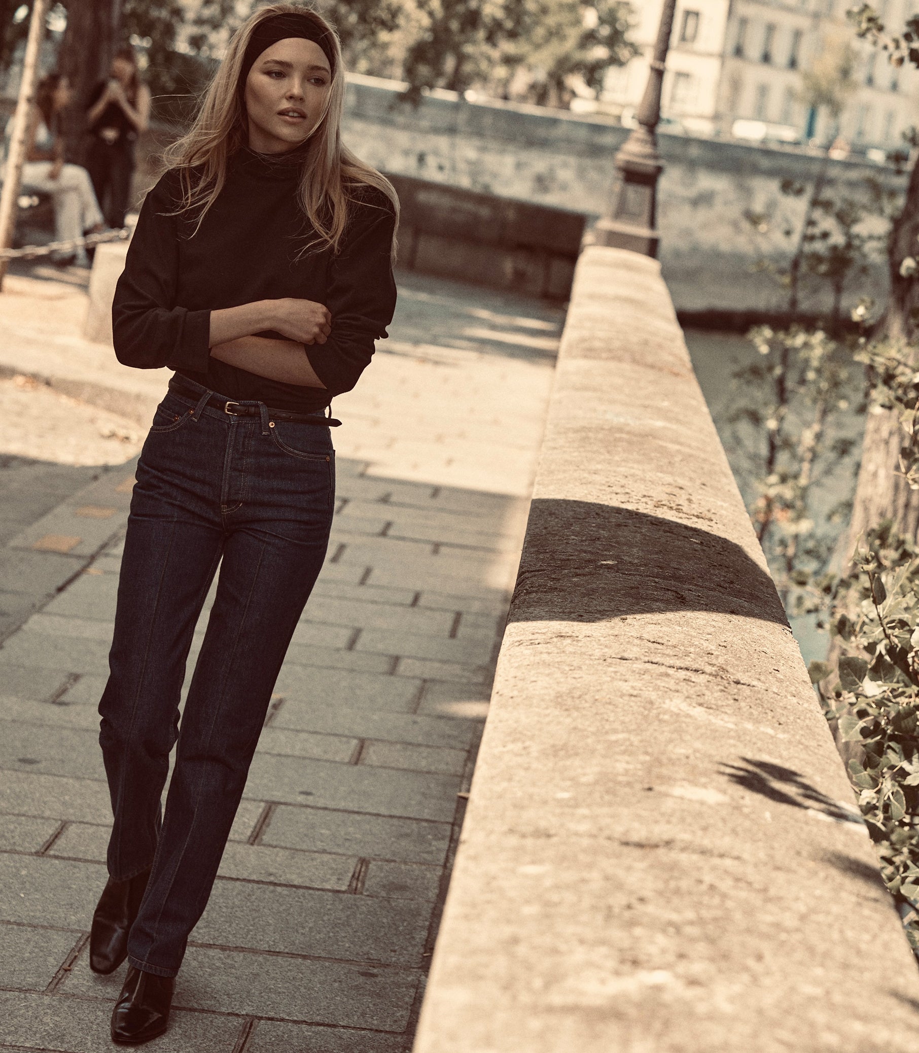 A woman wearing a black long-sleeve mock-neck top, dark blue jeans, and a black headband walks outdoors. 1
