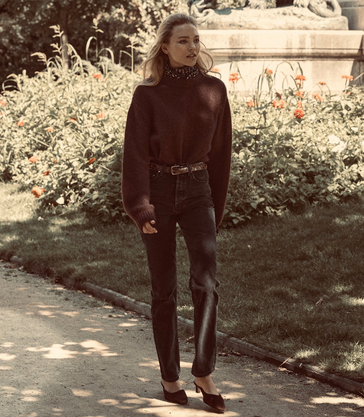 A woman stands outdoors in a park-like setting wearing a dark brown ribbed mock-neck sweater, dark wash jeans, and dark pointed-toe heels.