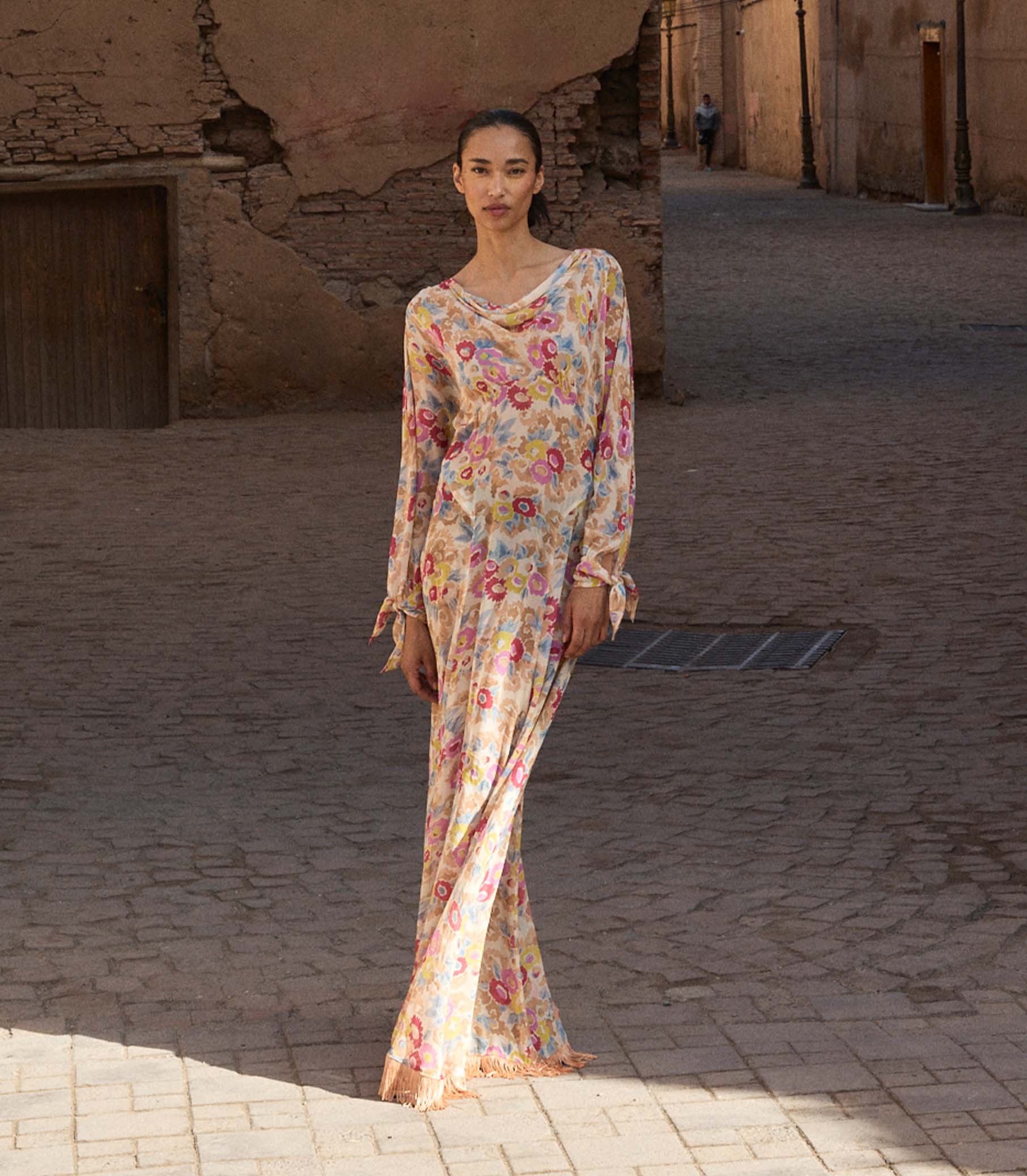 Woman in a long floral dress standing in a sunlit alleyway. 1
