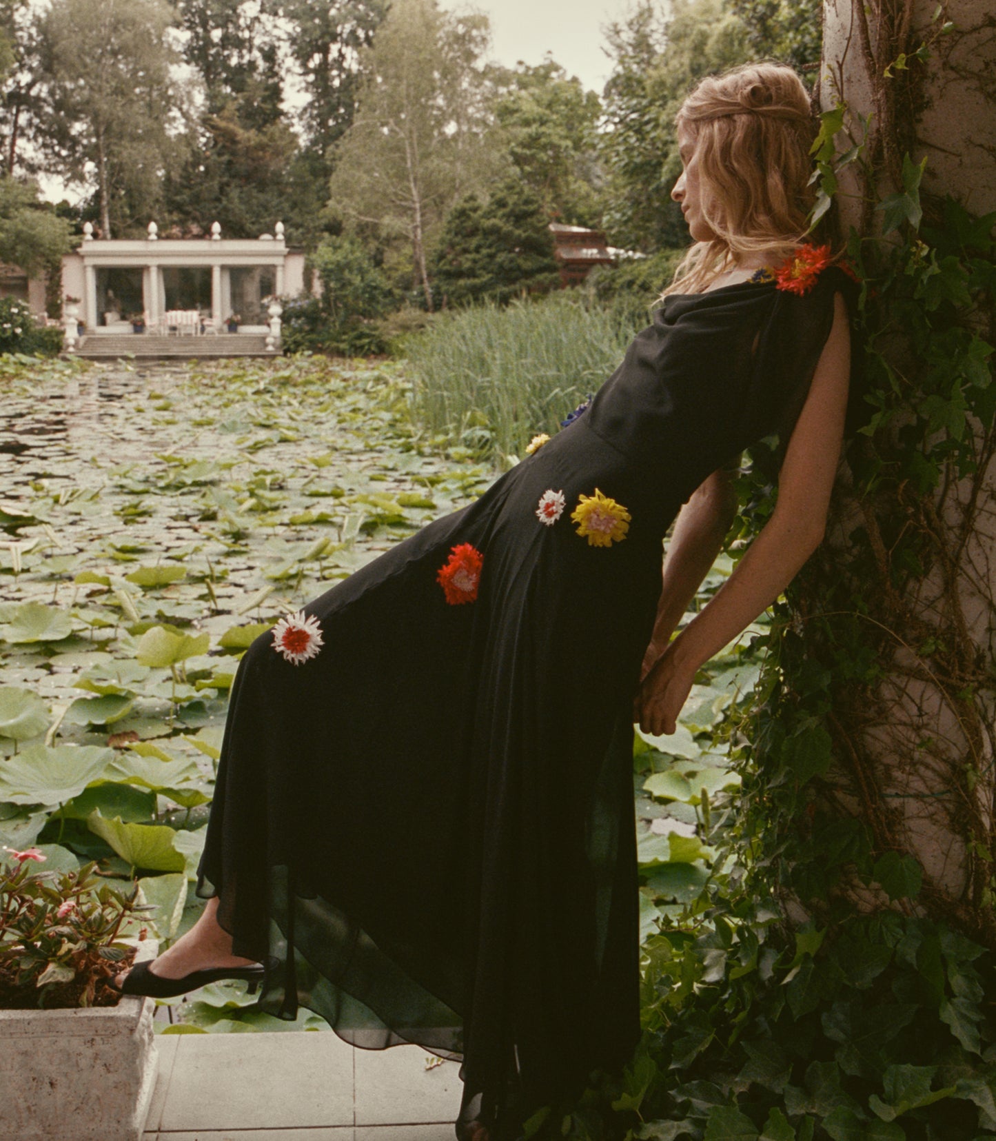 A woman in a black maxi dress with colorful flower appliqués leans against a vine-covered wall beside a lily pond.