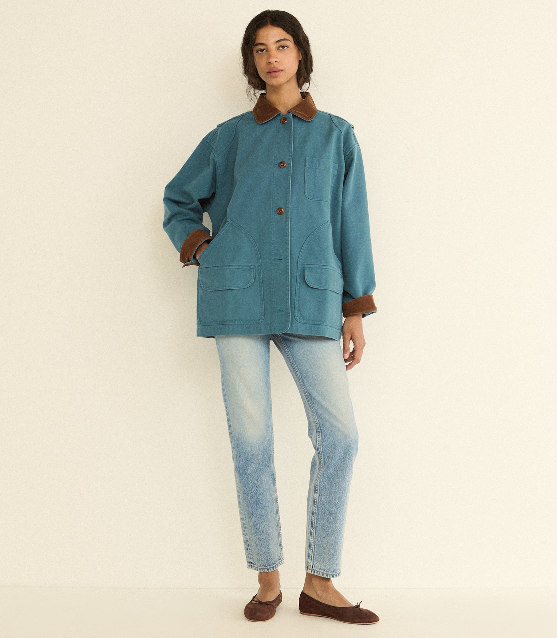 A model stands against a plain off-white studio background wearing a teal blue utility-style jacket with brown collar, cuffs, and button details, paired with light-wash jeans and brown flats.