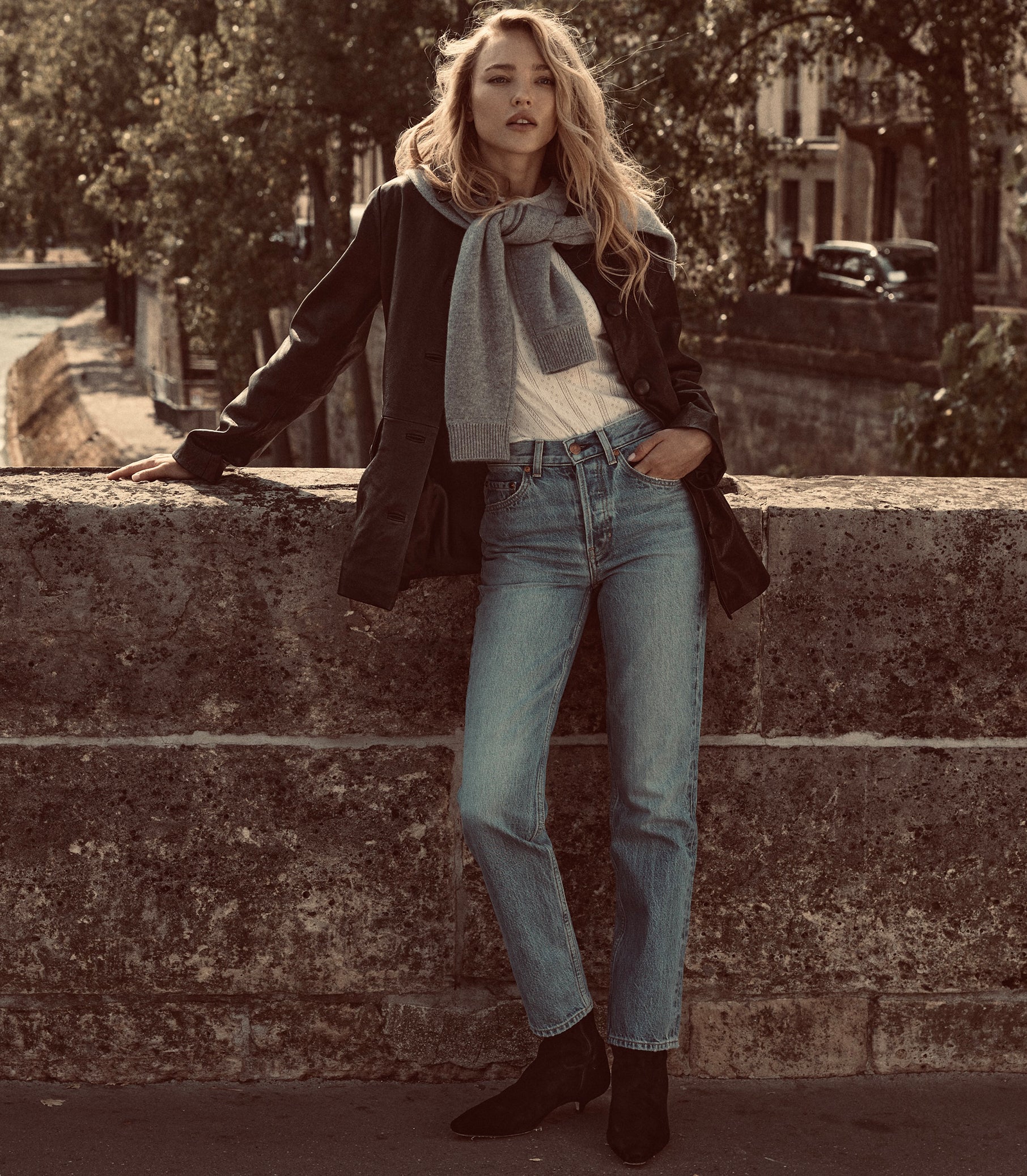 A woman stands outdoors leaning against a stone wall, wearing a black coat, light gray scarf, white top, light blue jeans, and black ankle boots. 1
