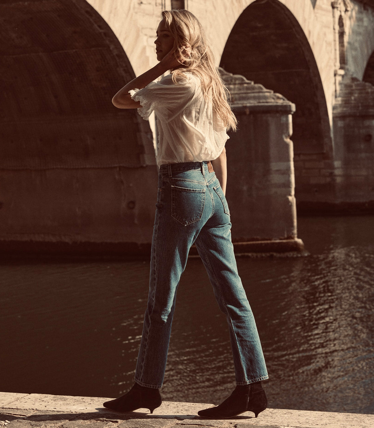 A woman stands by a river under a stone bridge, wearing high-waisted blue jeans, a white blouse, and black ankle boots.