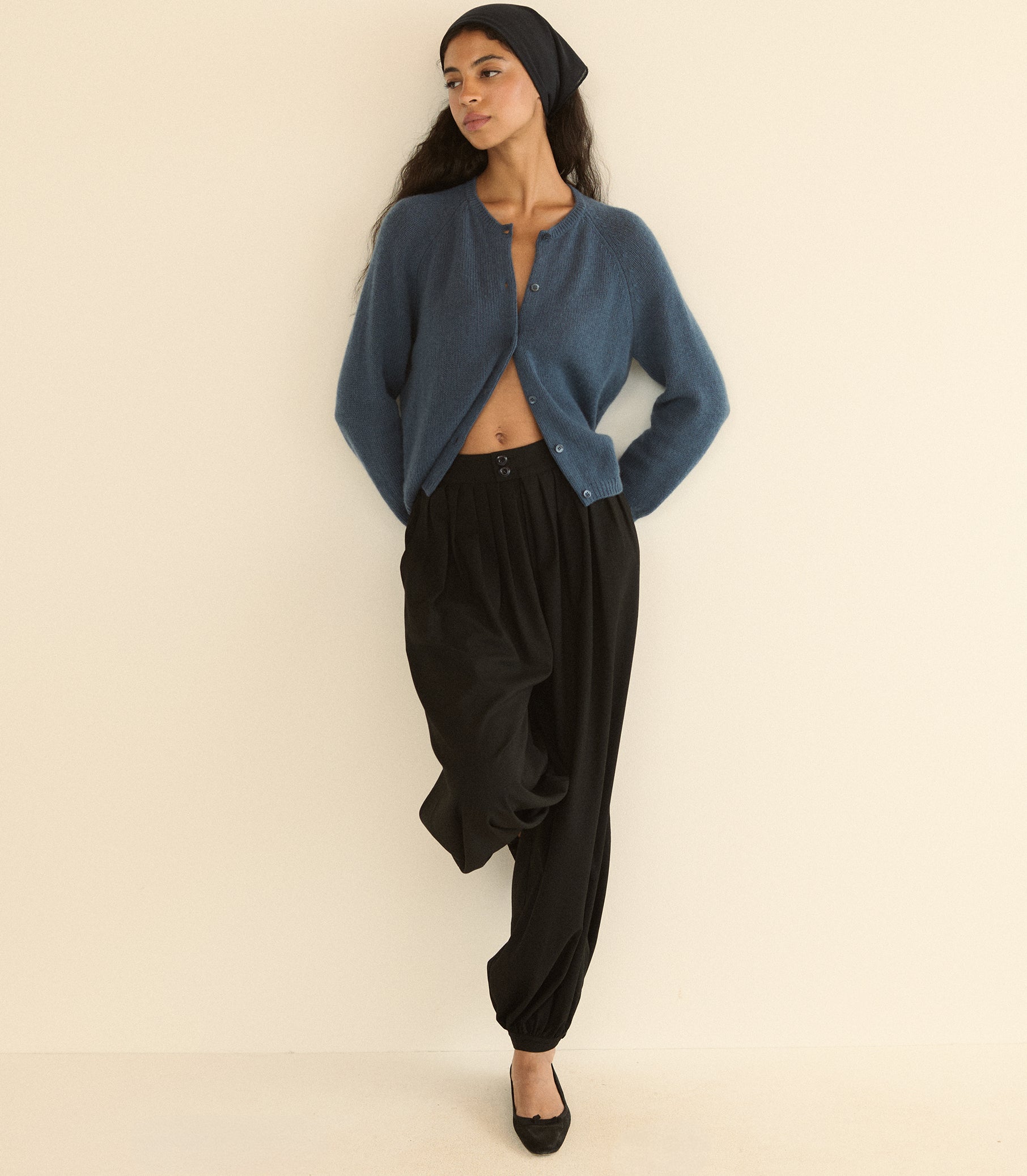 A woman modeling a blue cropped cardigan, black harem pants, a black beanie, and black flats against a plain cream background.