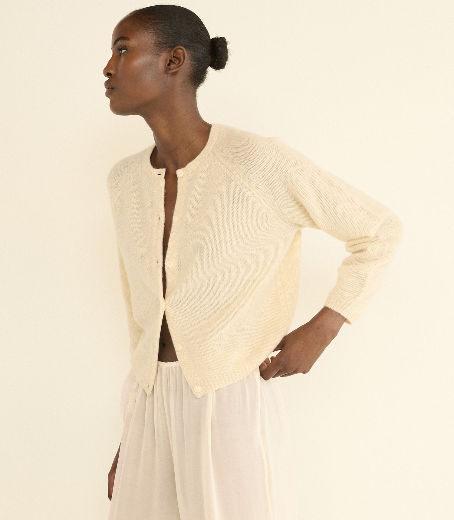 A model wearing a cream-colored, long-sleeve knit cardigan and cream trousers, shown from a side profile against a solid cream background.