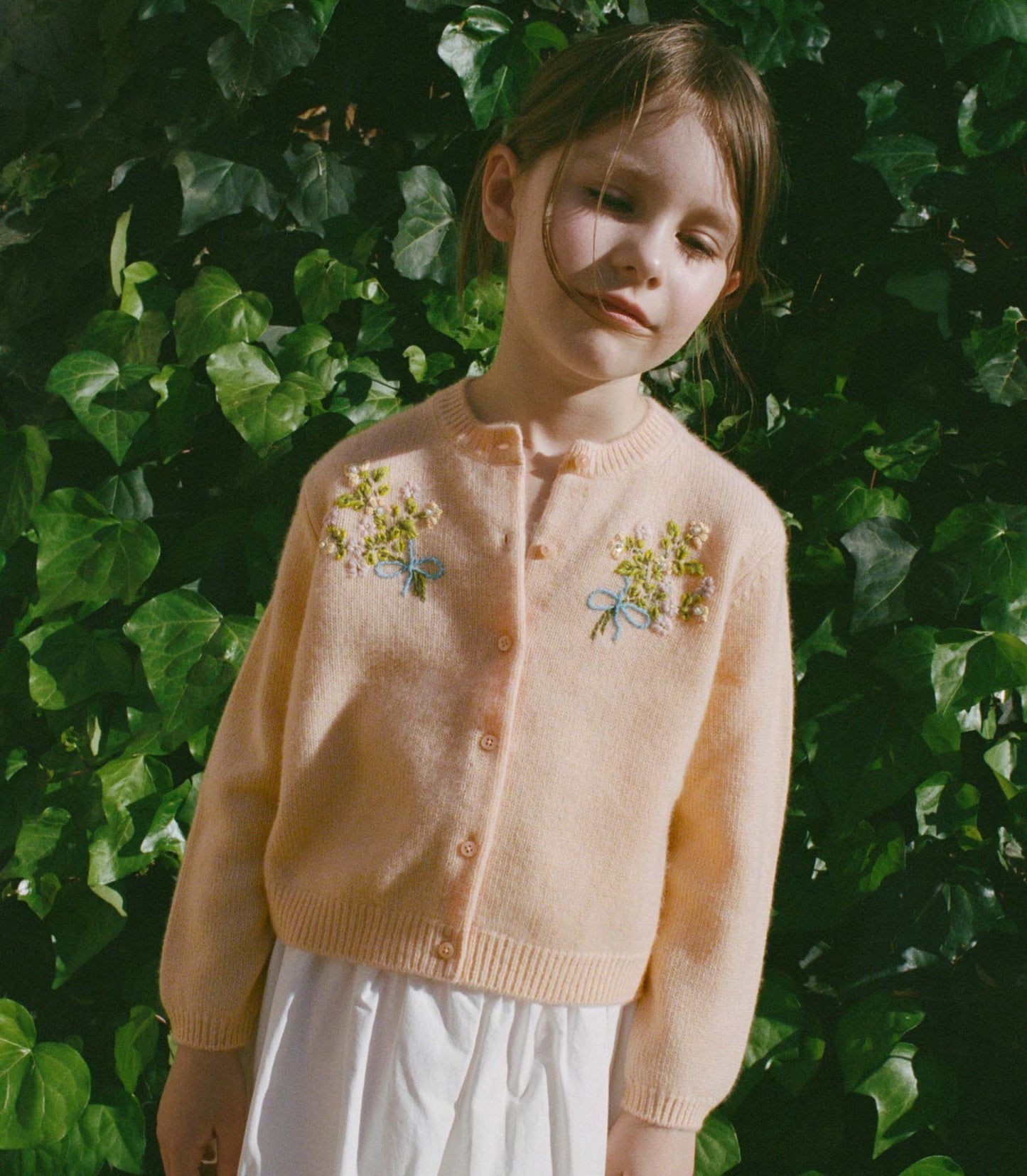 Young girl wearing a light pink cardigan with floral embroidery against a green leafy background