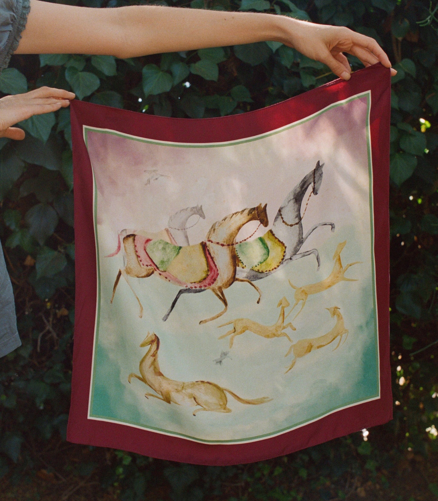 A square silk bandana with a deep red border and a central illustration of horses and gazelles, held up by a person's hands against a background of green foliage.