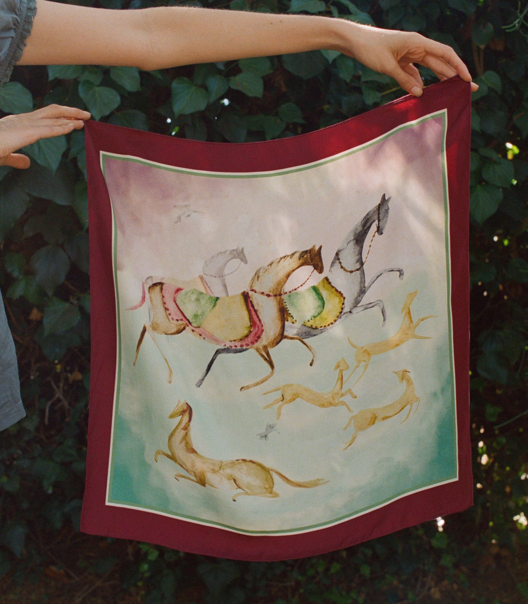A square silk bandana with a deep red border and a central illustration of horses and gazelles, held up by a person's hands against a background of green foliage.