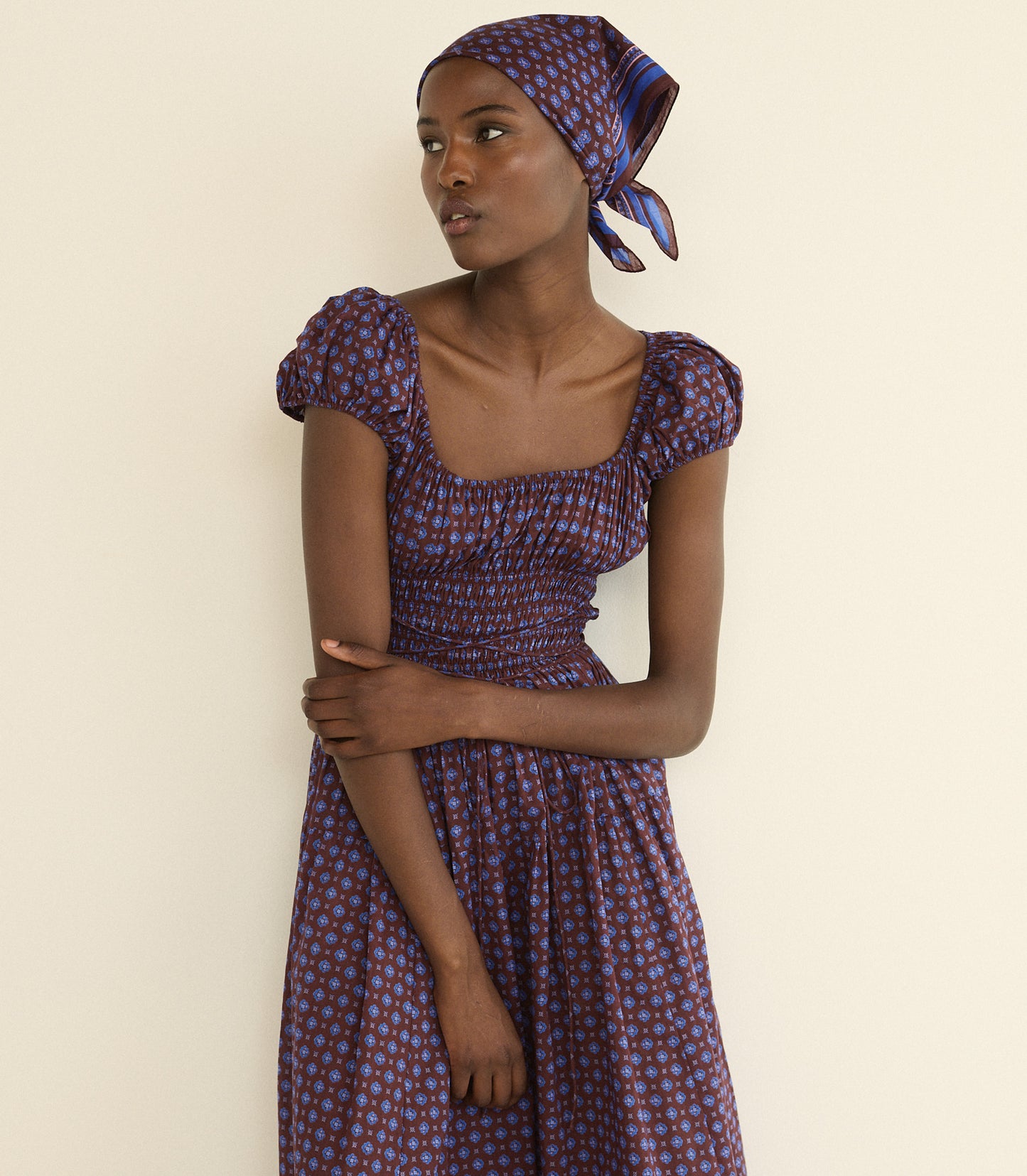 A woman wearing a purple and blue foulard bandana and a matching dress with puff sleeves, posed against a plain cream-colored wall.