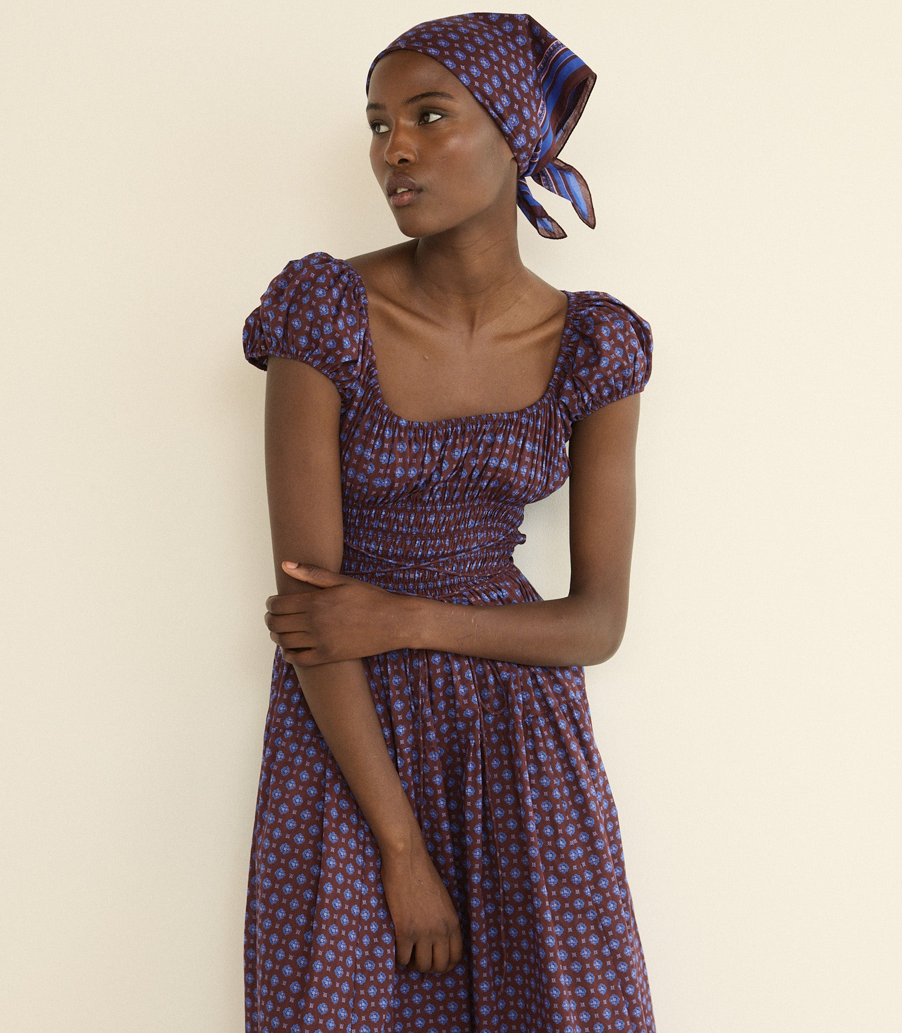 A woman wearing a purple and blue foulard bandana and a matching dress with puff sleeves, posed against a plain cream-colored wall. 1
