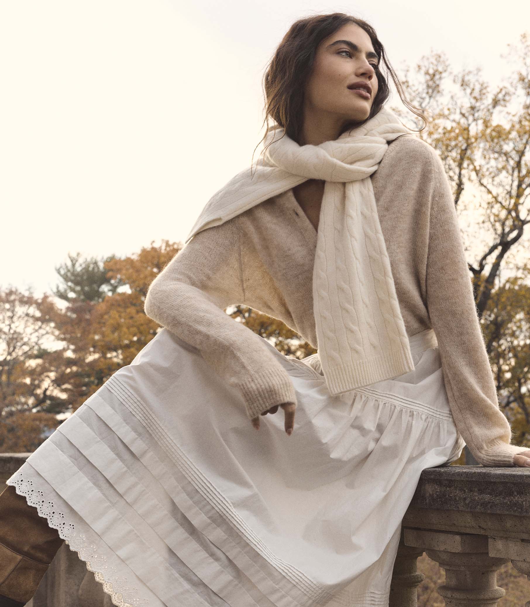 Woman wearing a beige sweater and scarf with a white dress, sitting outdoors. 2
