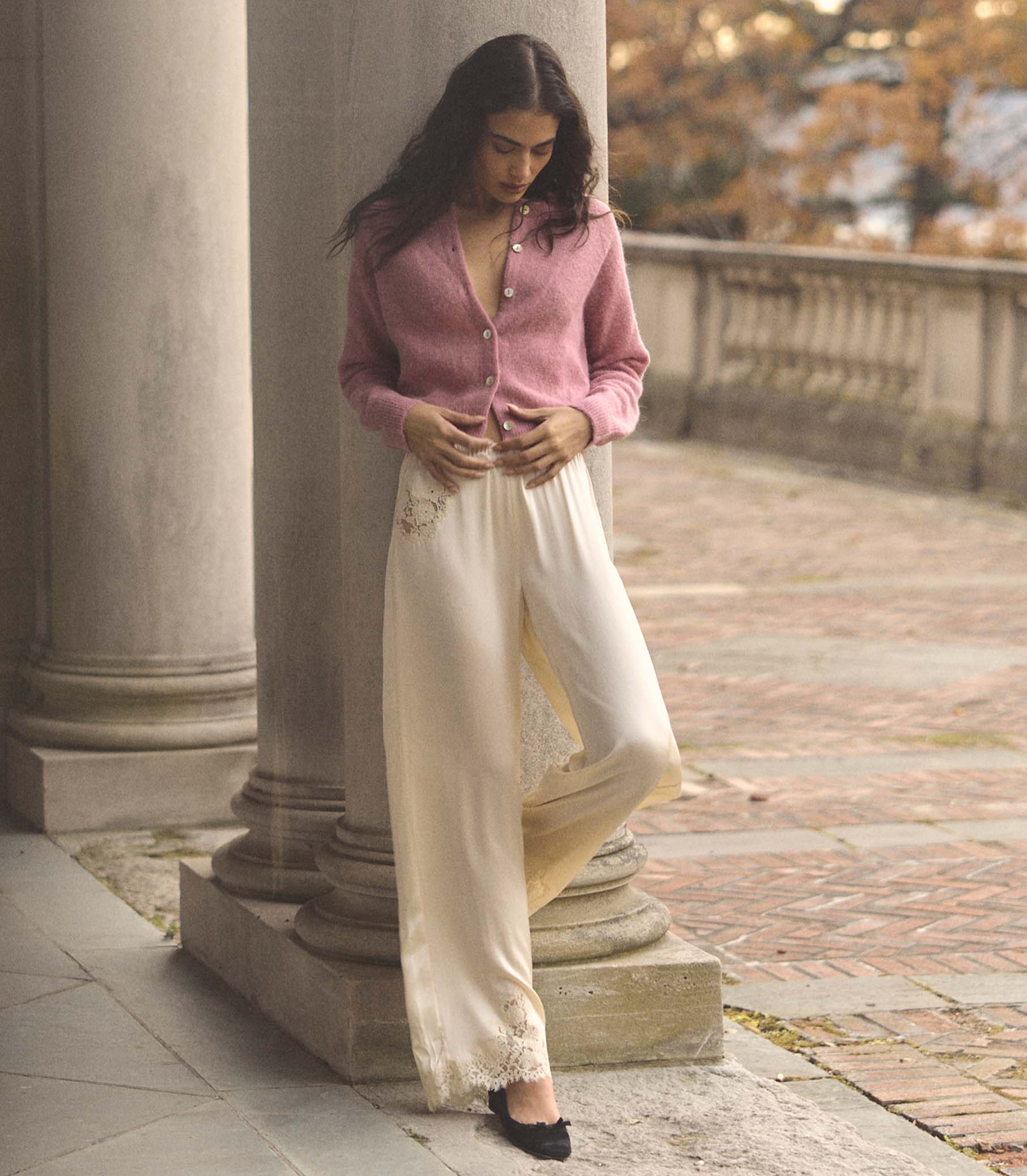 Woman in a pink cardigan and white pants standing on a stone path with columns in the background. 1
