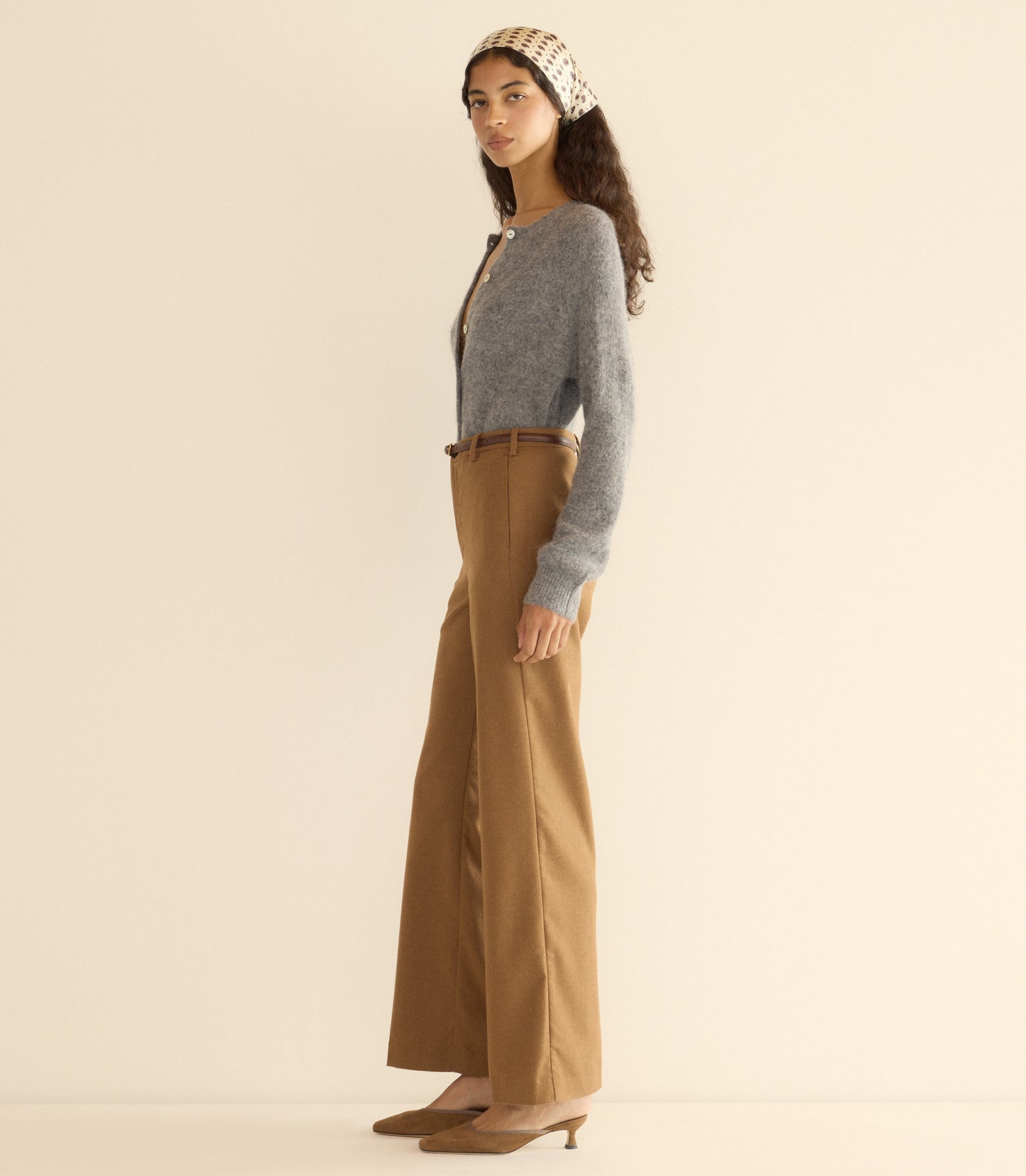 Woman wearing a gray cardigan and brown pants with a beige headband against a plain background