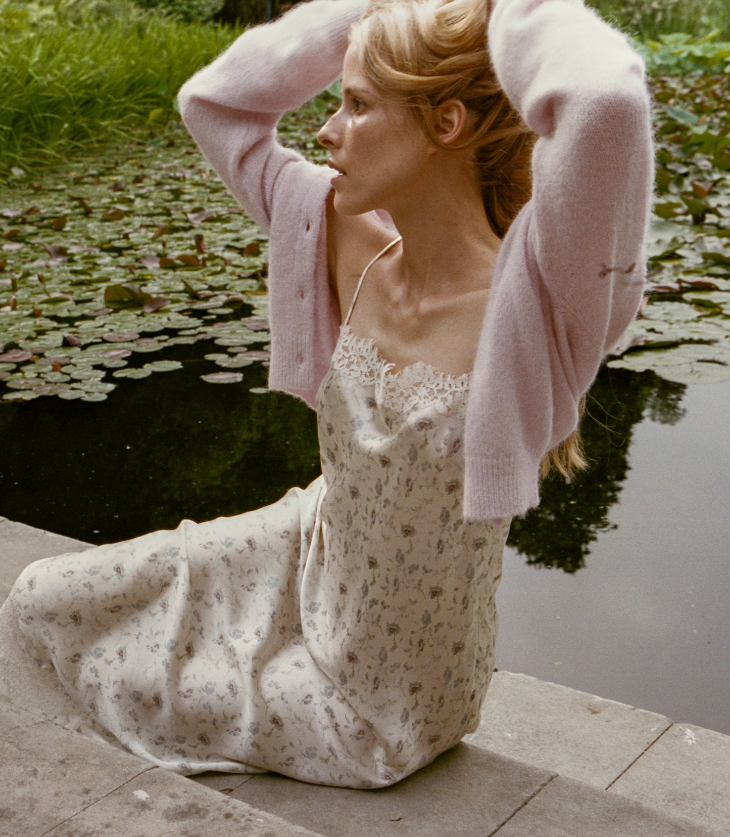 A woman sits by a pond wearing a cream floral slip dress with a light pink knit cardigan draped off her shoulders. She has her hands raised to her head, with lily pads and green grass visible in the background.