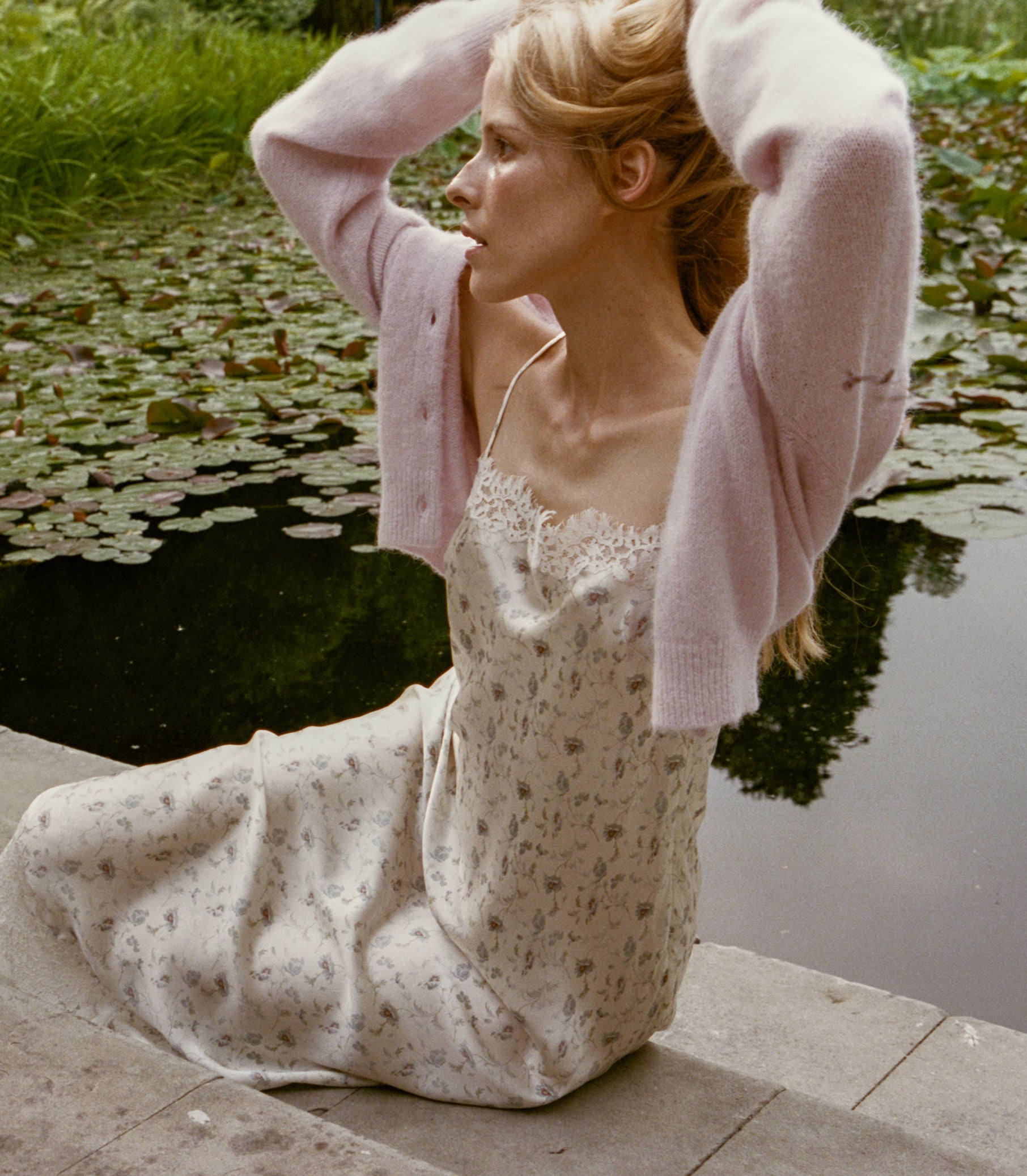 A woman sits by a pond wearing a cream floral slip dress with a light pink knit cardigan draped off her shoulders. She has her hands raised to her head, with lily pads and green grass visible in the background. 1
