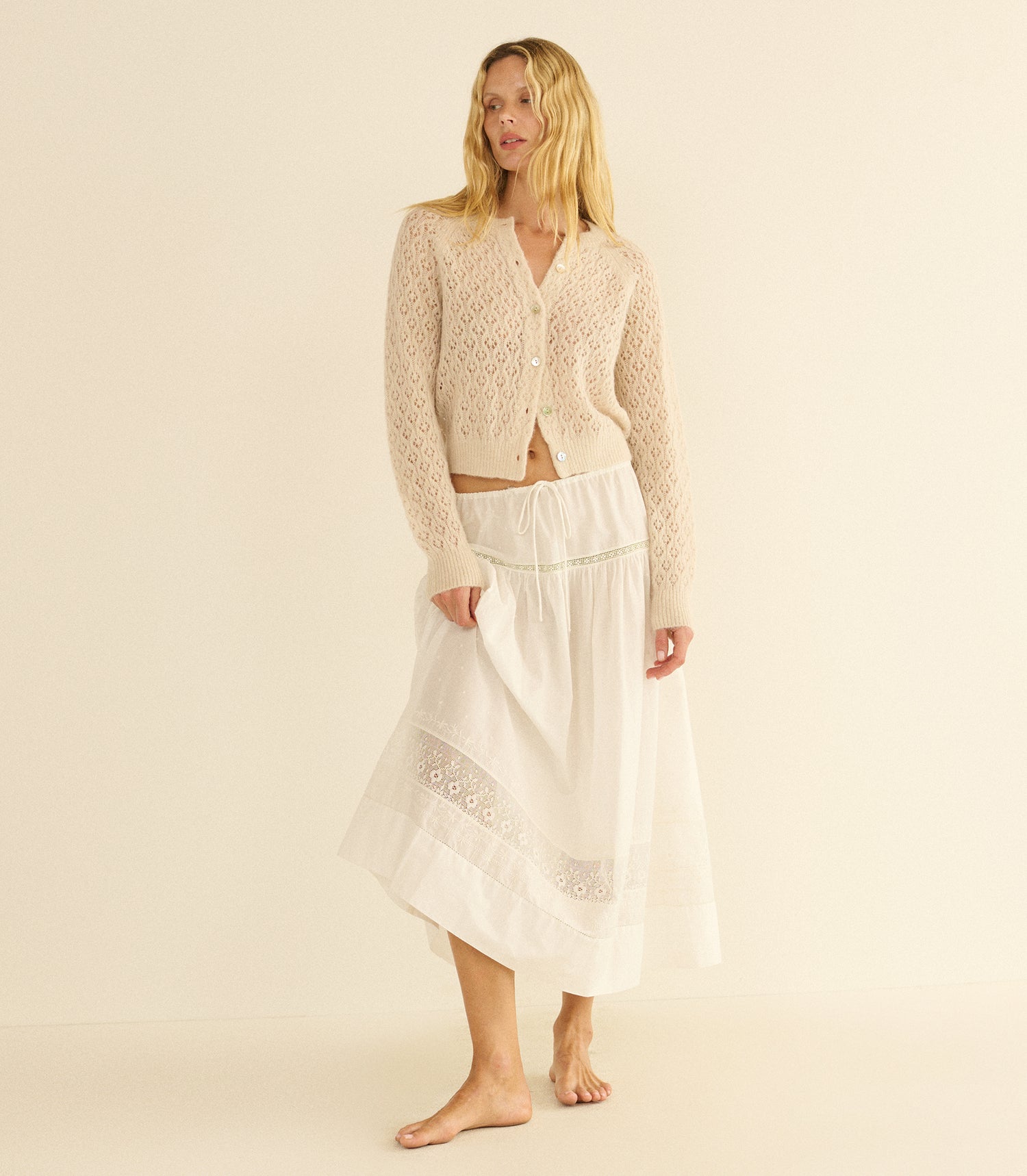Woman wearing a beige pointelle cardigan and white skirt on a beige background