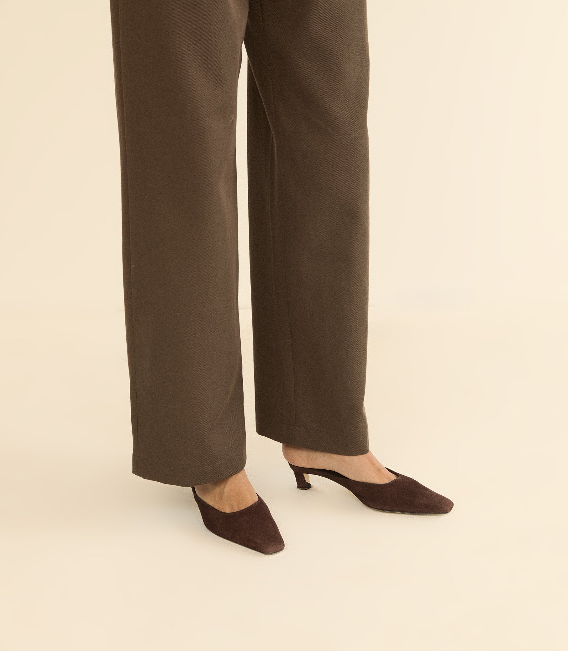 A person wearing dark brown wide-leg trousers and matching pointed-toe mule heels, shown from the knees down against a plain warm beige studio background. 1
