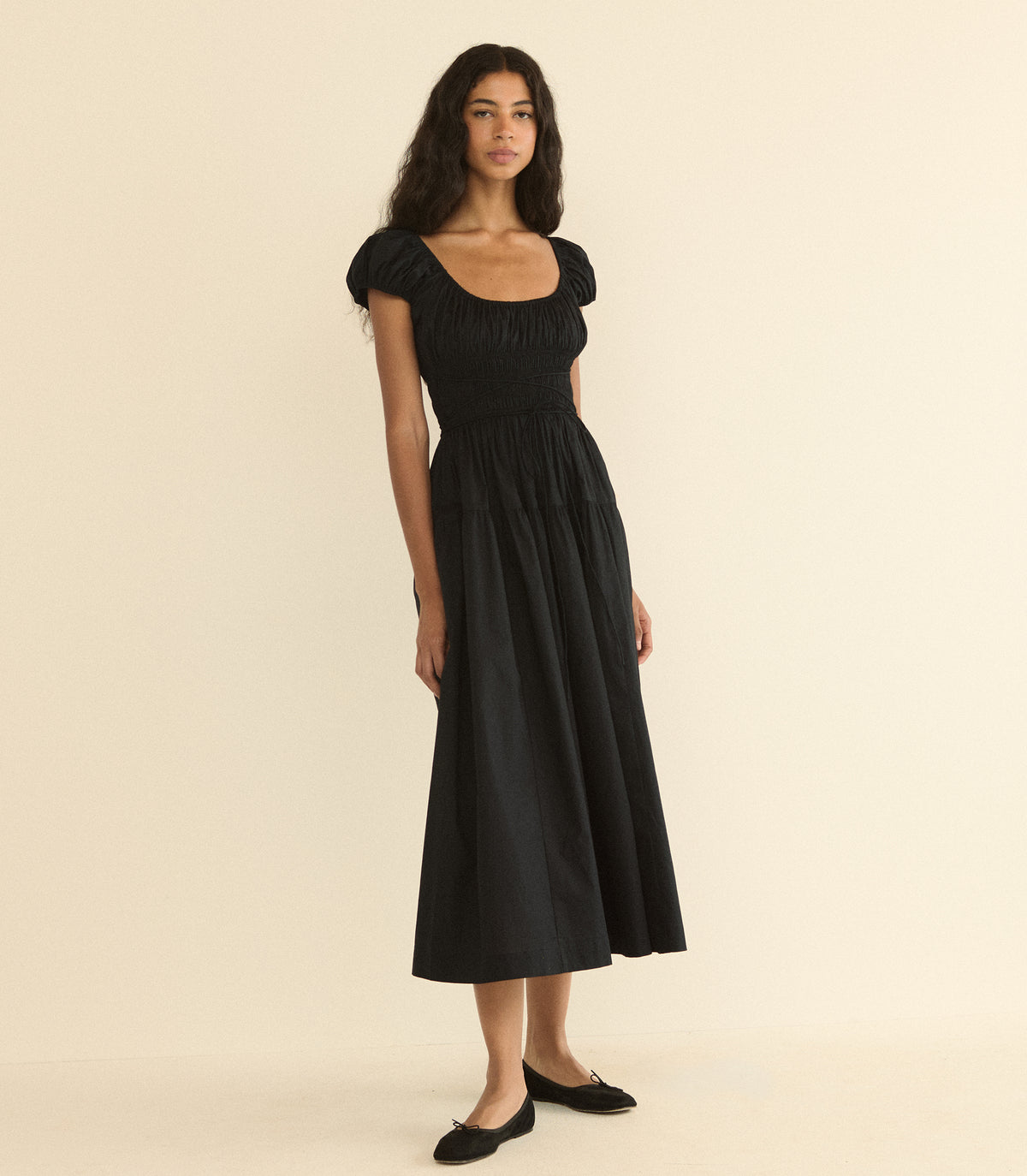 A woman modeling a black midi dress with a smocked bodice, puffed cap sleeves, and a tiered skirt, paired with black flats, against a solid cream background.