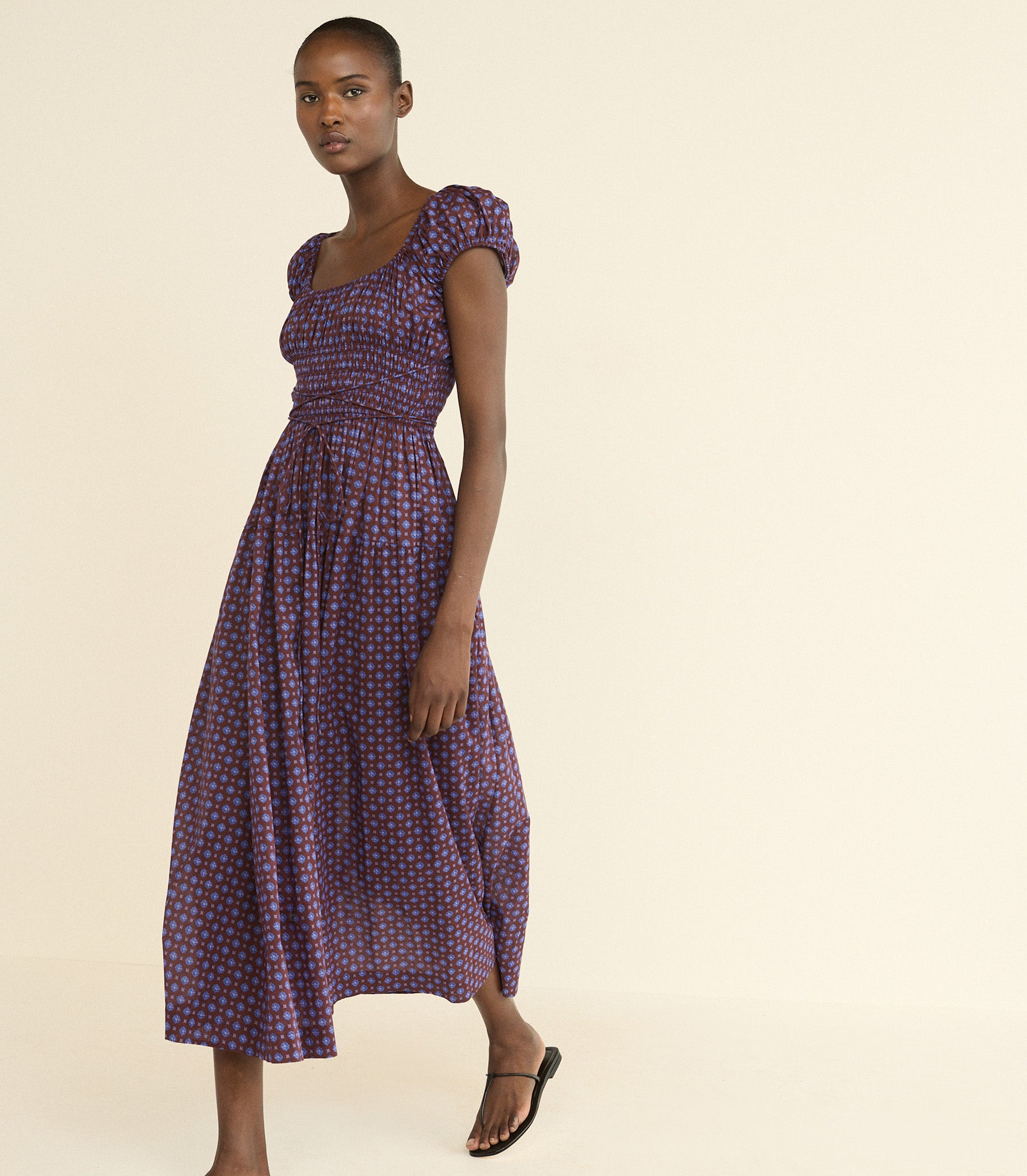 A woman modeling a midi-length purple dress with a small red and white foulard pattern, puffed cap sleeves, and a smocked bodice, set against a solid cream background. 1
