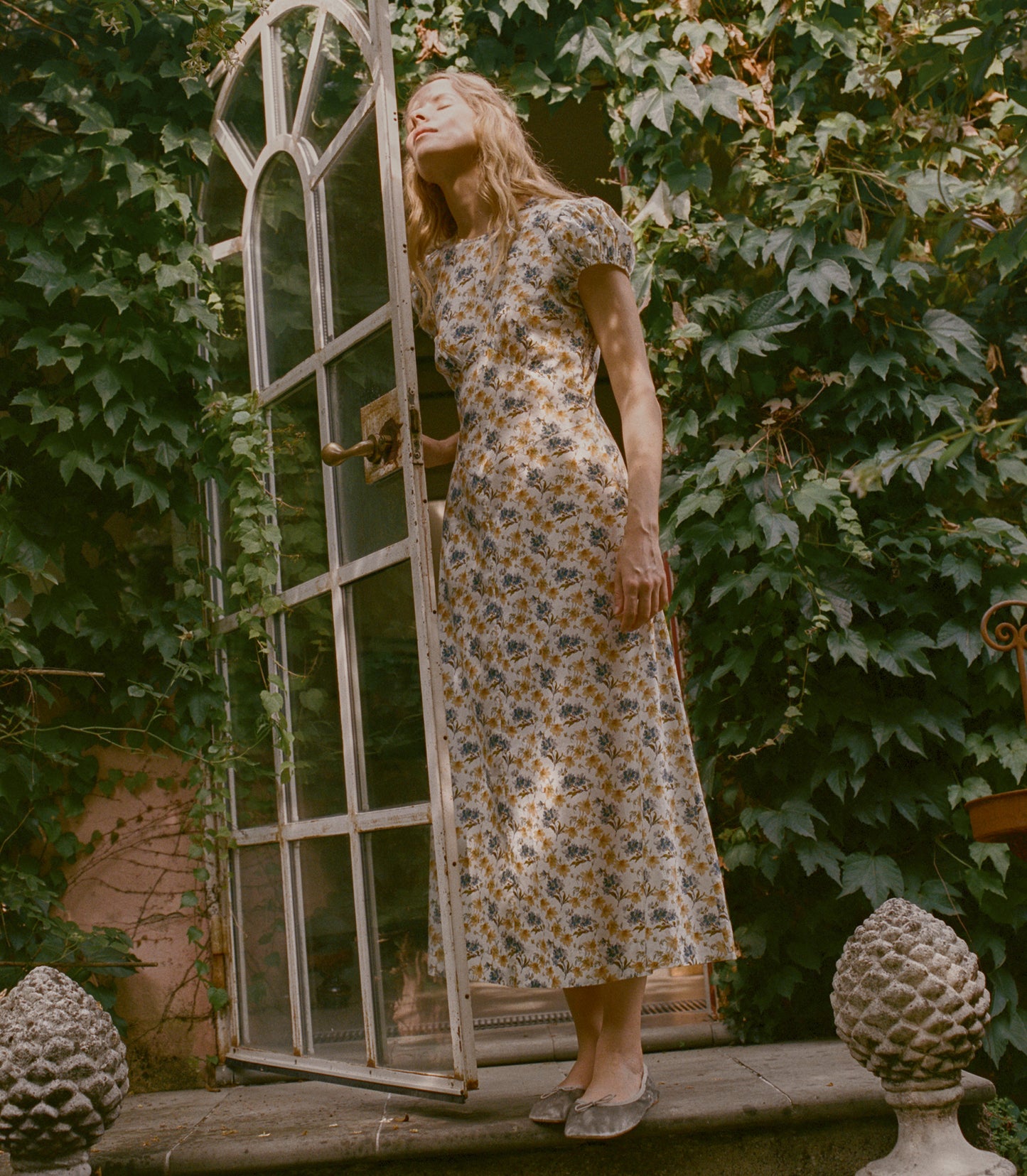 A woman wearing a short-sleeved midi dress with a blue and yellow floral pattern on a cream background, standing next to an open white arched window frame in a garden setting with ivy.
