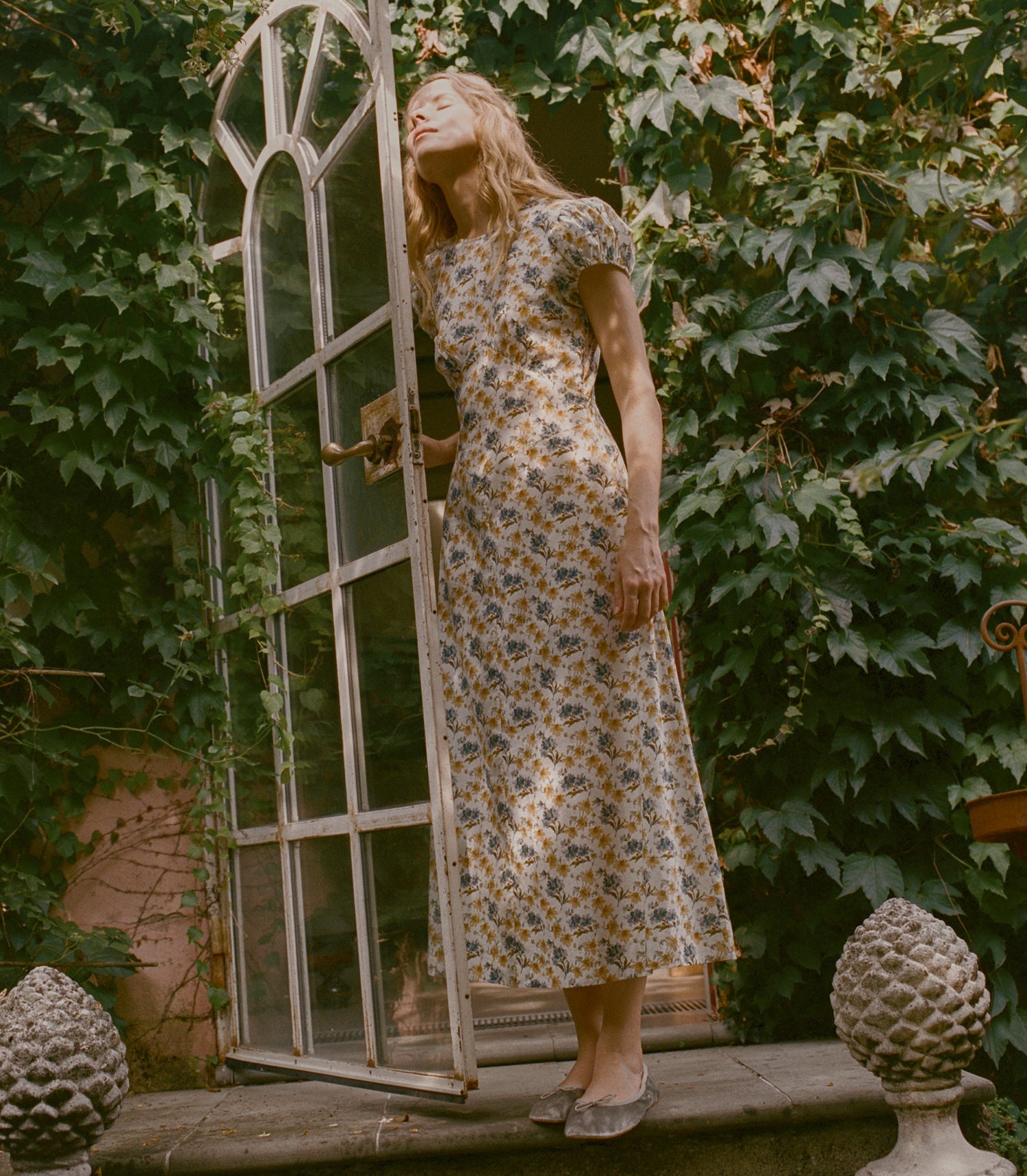 A woman wearing a short-sleeved midi dress with a blue and yellow floral pattern on a cream background, standing next to an open white arched window frame in a garden setting with ivy.