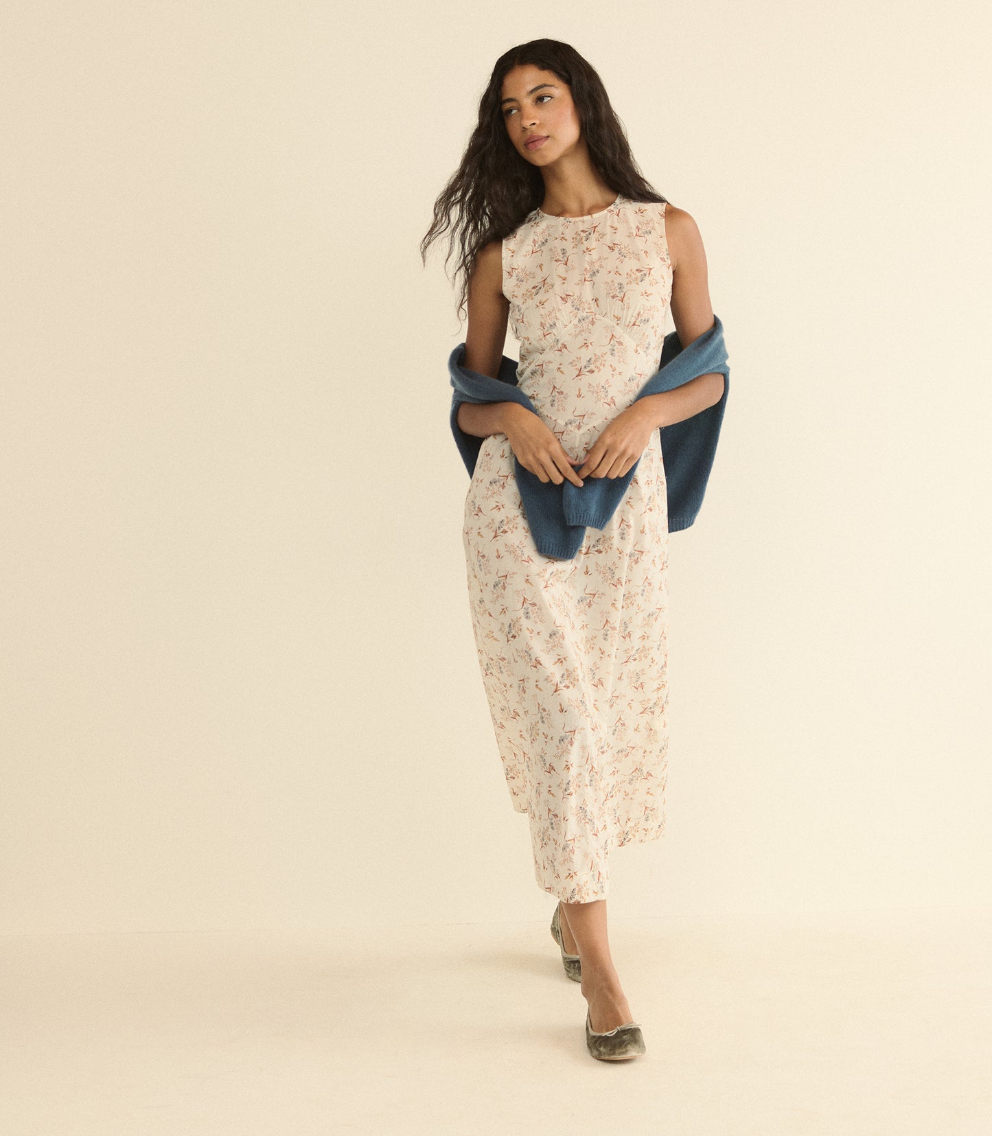 A woman modeling a sleeveless cream dress with a delicate floral print, styled with a draped blue shawl and metallic flat shoes, set against a solid warm beige studio background.