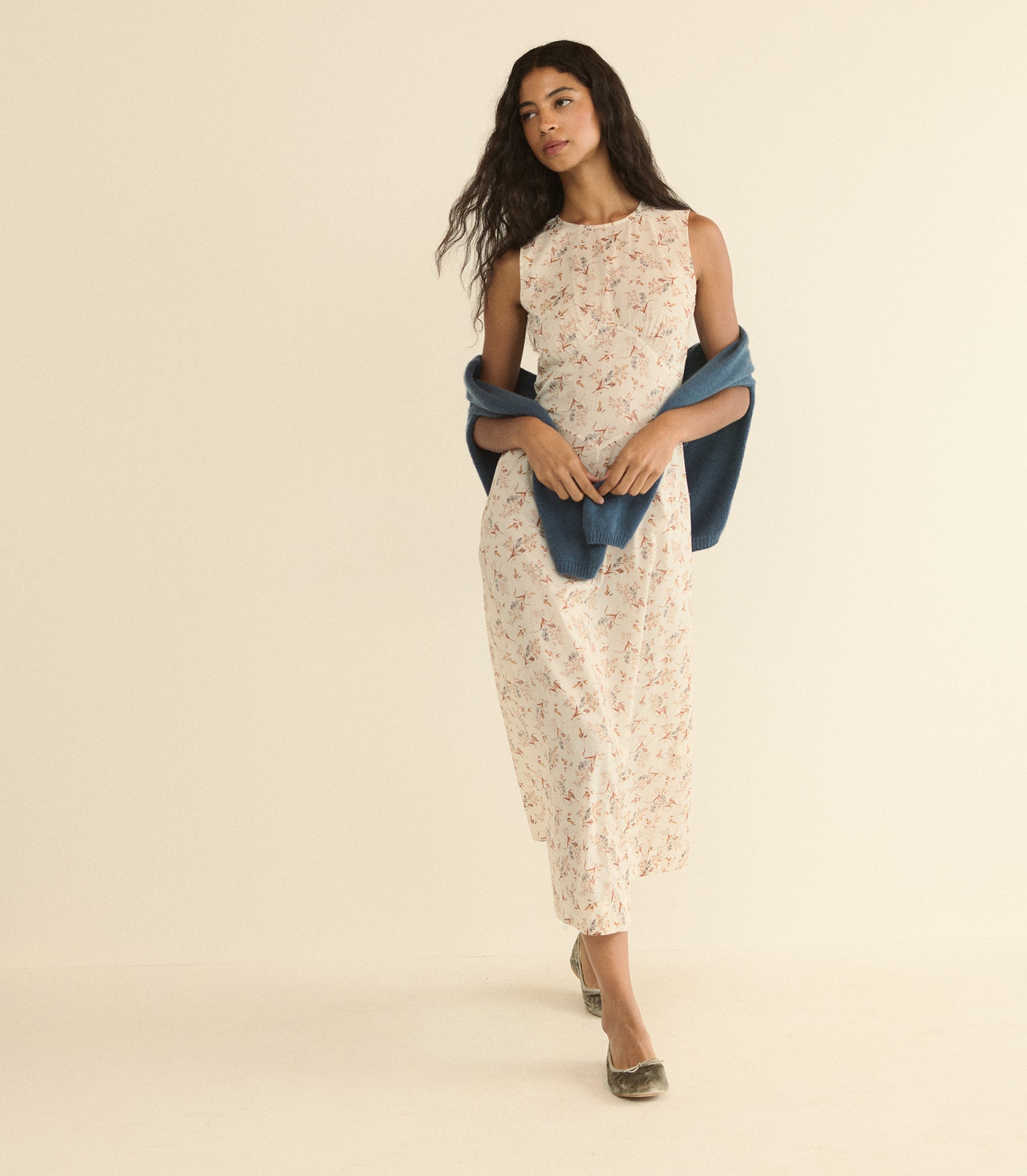 A woman modeling a sleeveless cream dress with a delicate floral print, styled with a draped blue shawl and metallic flat shoes, set against a solid warm beige studio background. 1
