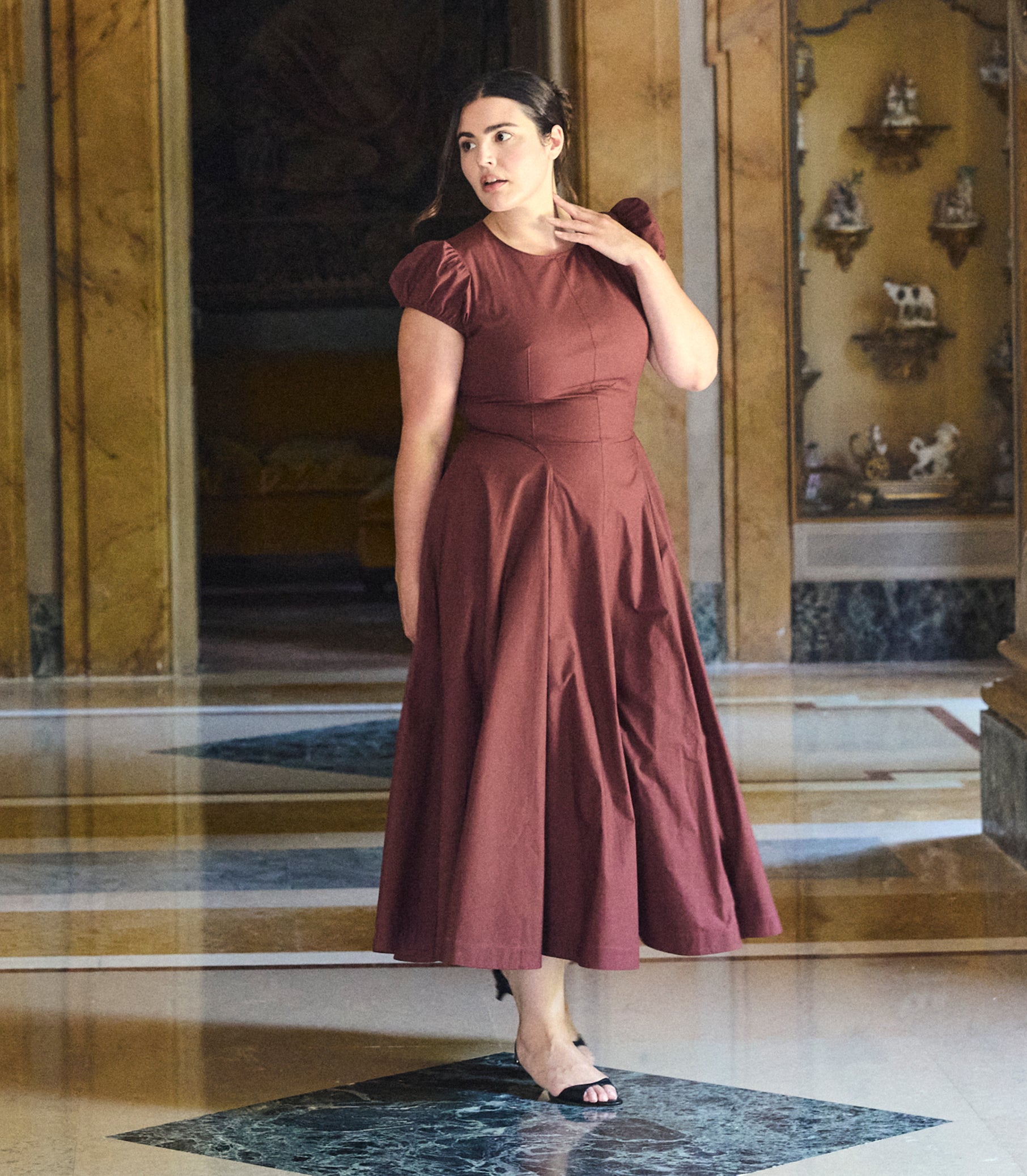A woman wearing a rust-brown midi dress with short puff sleeves and a gathered waist, standing in a grand interior hallway with marble floors and gold-toned walls.