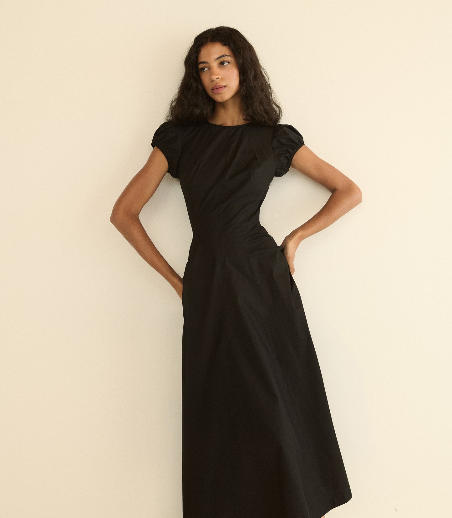 A woman modeling a black maxi dress with short puff sleeves, a round neckline, and a gathered waist, posing against a plain off-white background.