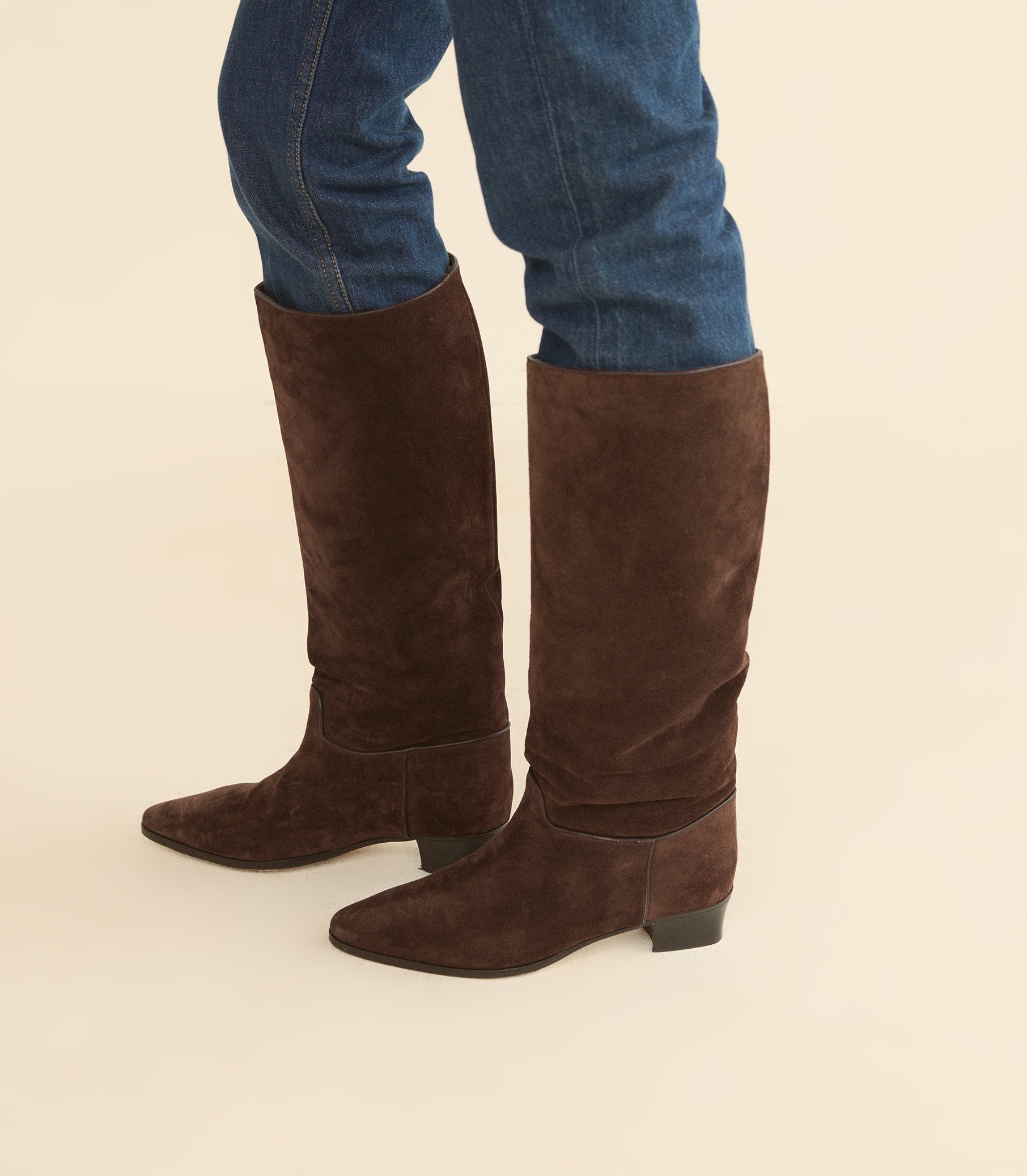 A pair of dark brown suede knee-high boots worn with blue jeans, shown from the knees down against a solid light beige background. 1
