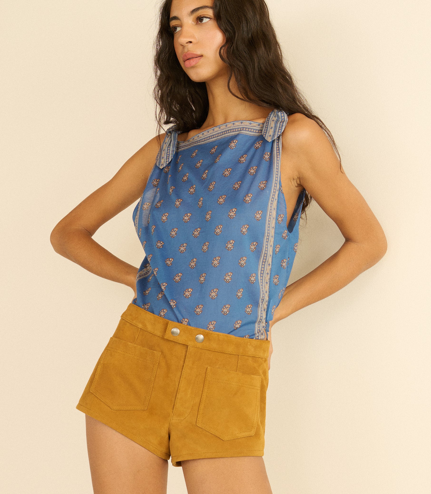 Woman wearing a blue paisley top and mustard yellow suede shorts against a beige background