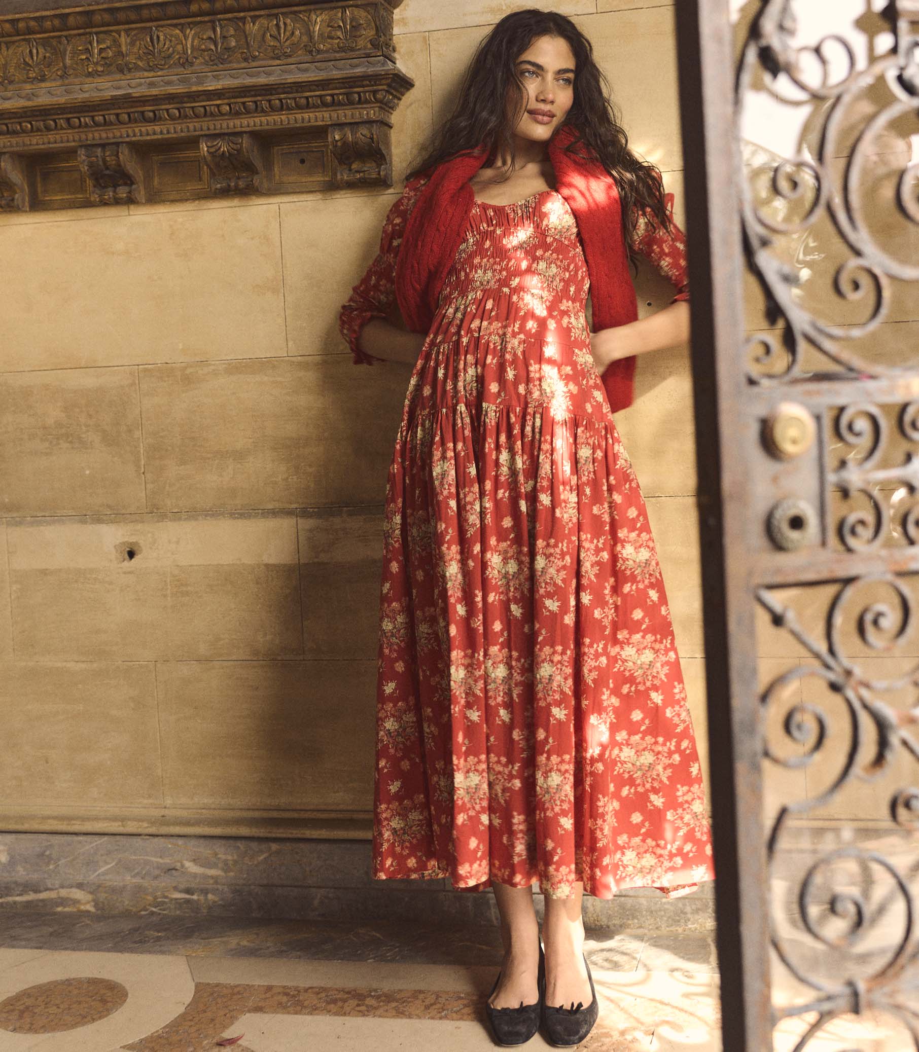 Woman wearing a red floral dress standing in a decorative interior setting. 1
